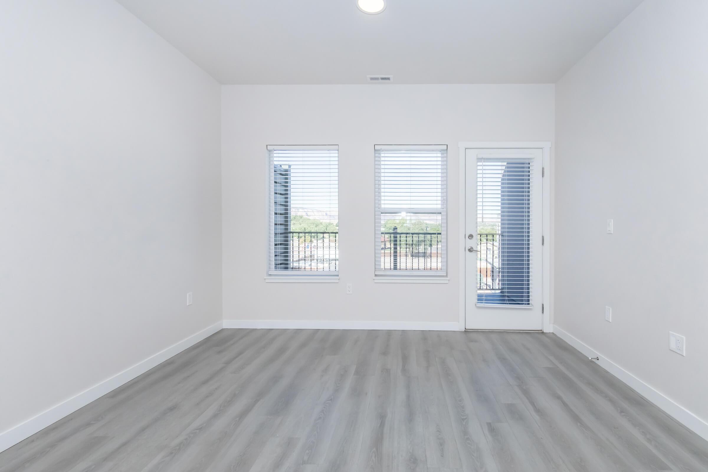 Empty room with light gray walls and a light gray wood-like floor. Two windows with blinds allow natural light into the space, and a door leads to a small balcony. The room is unfurnished, providing a blank canvas for decoration.
