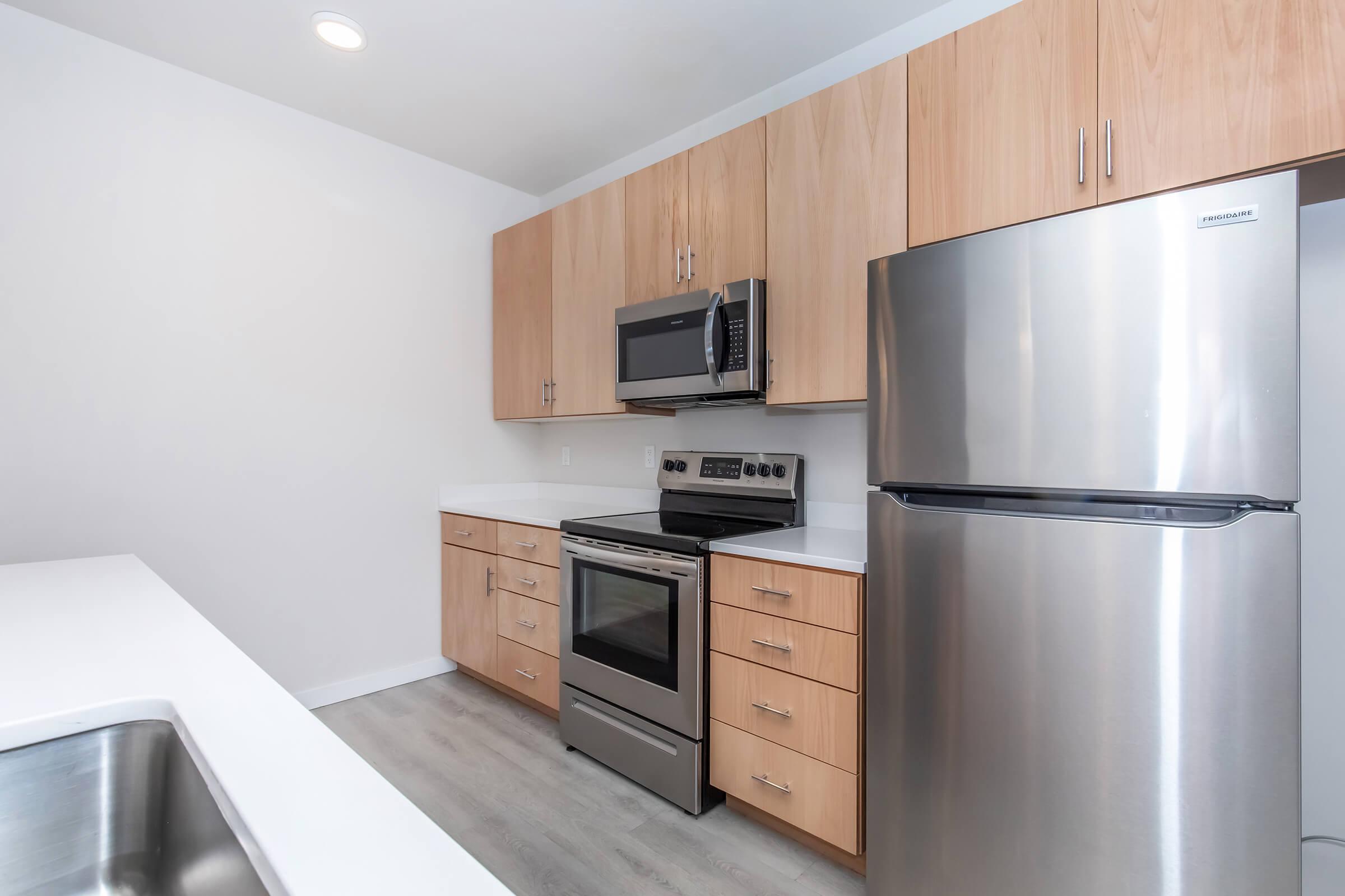 Modern kitchen with light wood cabinets, stainless steel appliances including a microwave, oven, and refrigerator. The countertop is white, and the flooring is light gray. The design is sleek and contemporary, featuring an open layout that enhances the spacious feel of the kitchen. 