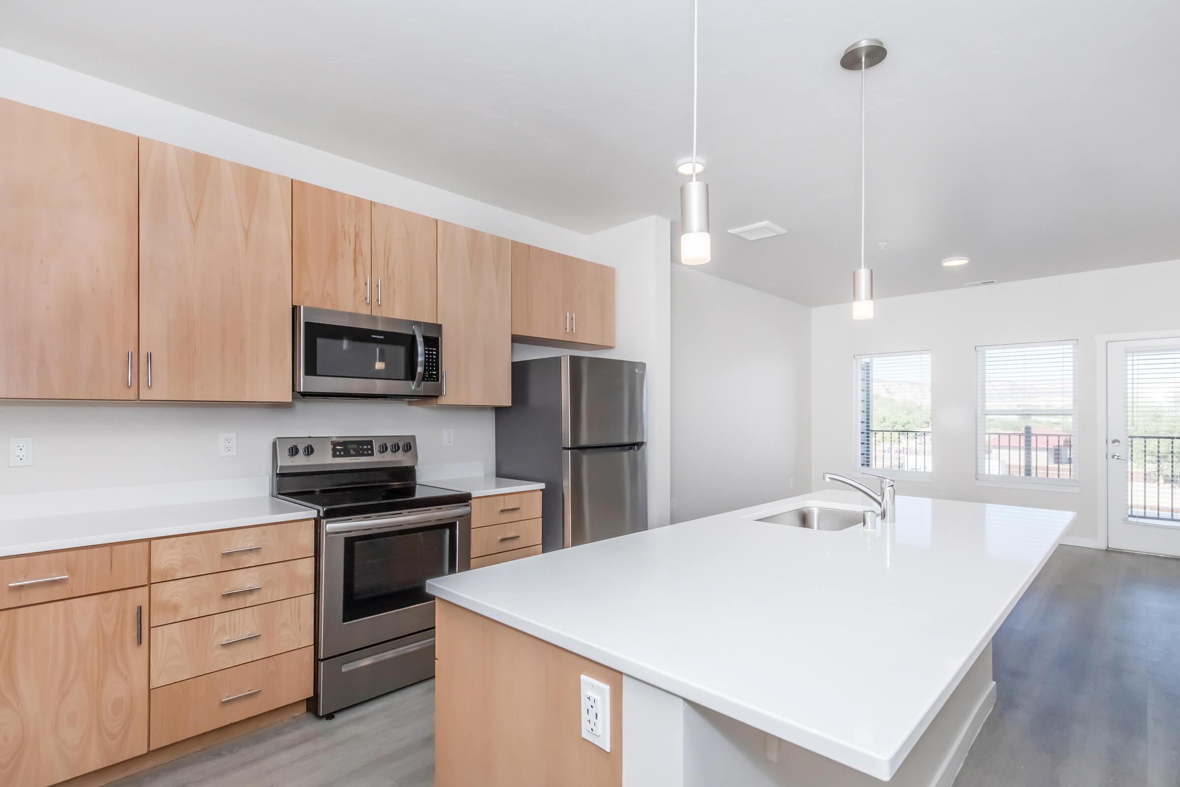Modern kitchen featuring light wood cabinetry, stainless steel appliances, including a microwave and oven, and a spacious white countertop island. Large windows provide natural light, and the open layout includes a dining area with contemporary fixtures.