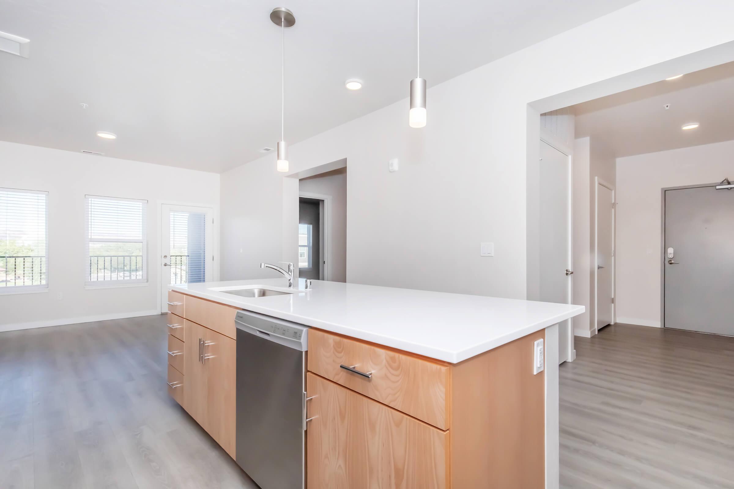 Modern kitchen featuring a large island with a built-in sink and stainless steel dishwasher. The cabinetry is light wood, and the countertops are white. Bright, airy space with large windows providing natural light. The flooring is a light-colored wood laminate, and there is a doorway leading to an adjacent room.