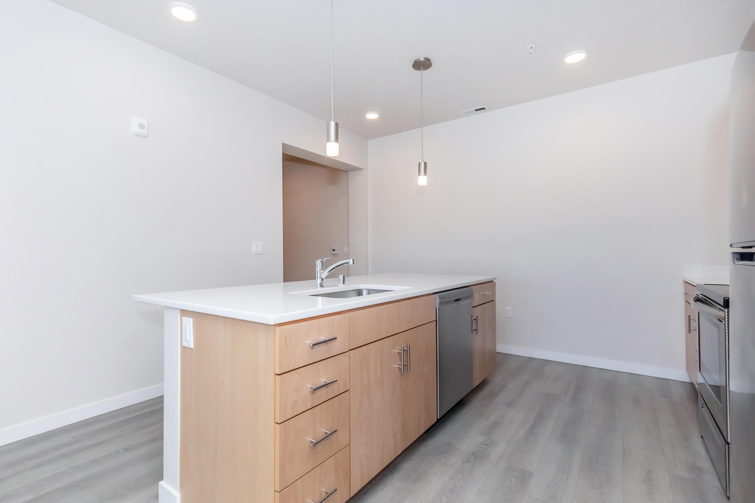 Modern kitchen featuring a central island with a sink, light wood cabinetry, stainless steel appliances, and pendant lighting. The space has a sleek, minimalistic design with light gray flooring and neutral-colored walls, creating a bright and open atmosphere.