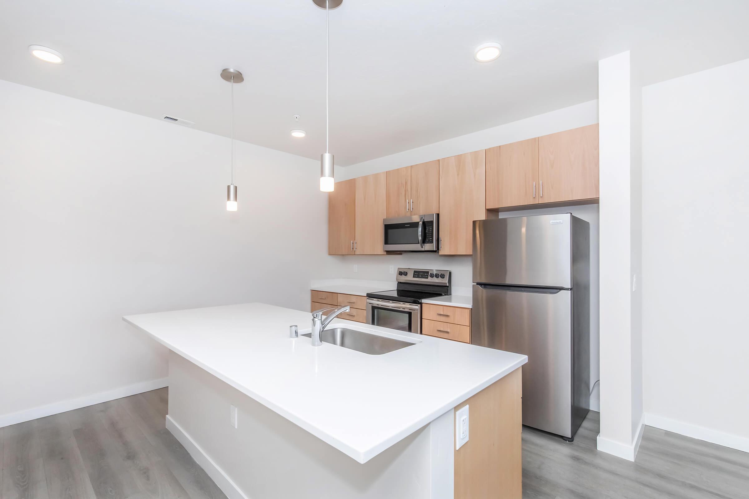 Modern kitchen featuring an island with a sink, stainless steel appliances including a microwave, oven, and refrigerator. Light wood cabinetry contrasts with a white countertop. The space has neutral-colored walls and subtle lighting fixtures, creating a bright and airy atmosphere.