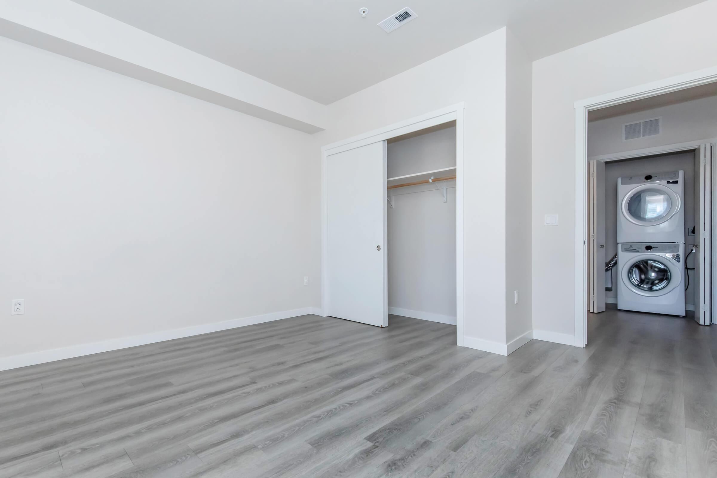 A bright, empty room with light-colored walls and light gray flooring. A closet with sliding doors is on the left, and a doorway leads to a laundry area featuring washing and drying machines in the background. The space is uncluttered and has ample natural light.