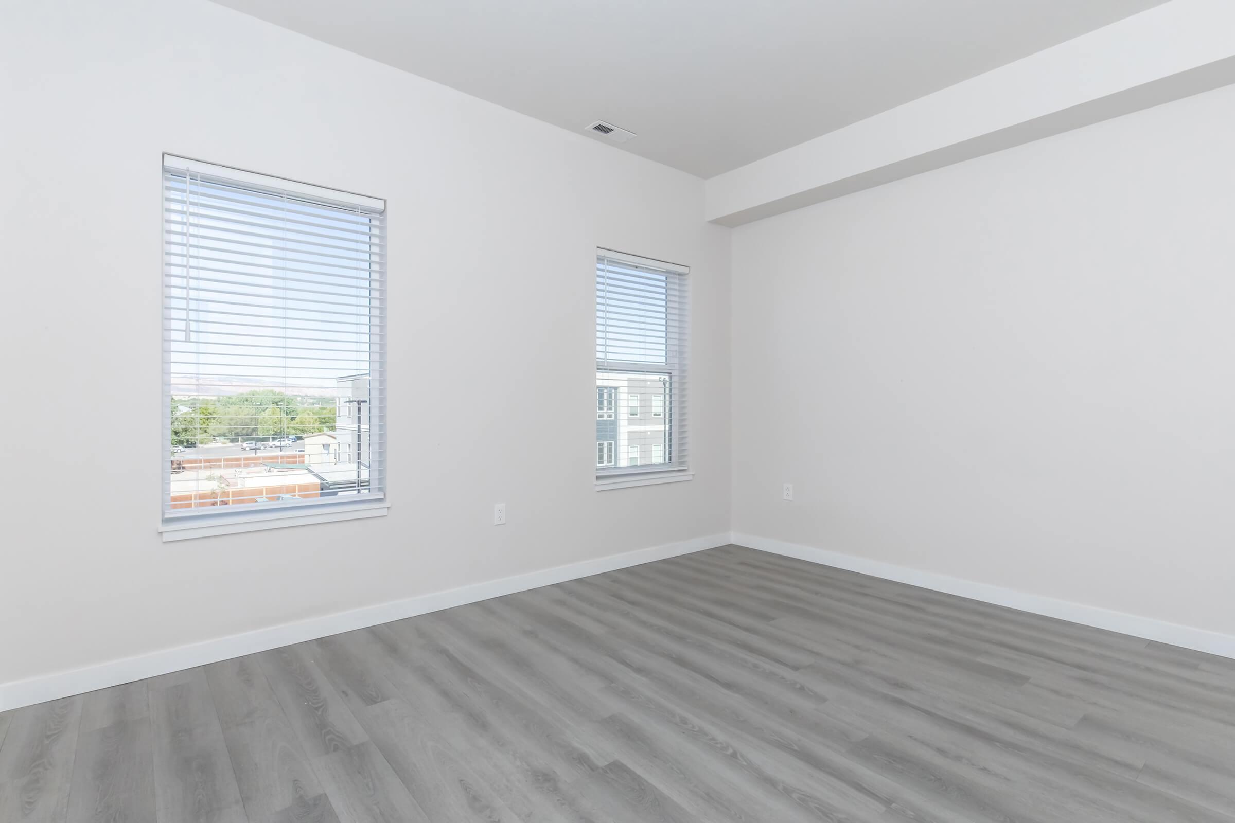 Spacious, empty room with light gray wood-like flooring, featuring two windows with white blinds. The walls are painted in a soft neutral color, allowing natural light to brighten the space. Suitable for various interior designs or furnishings.