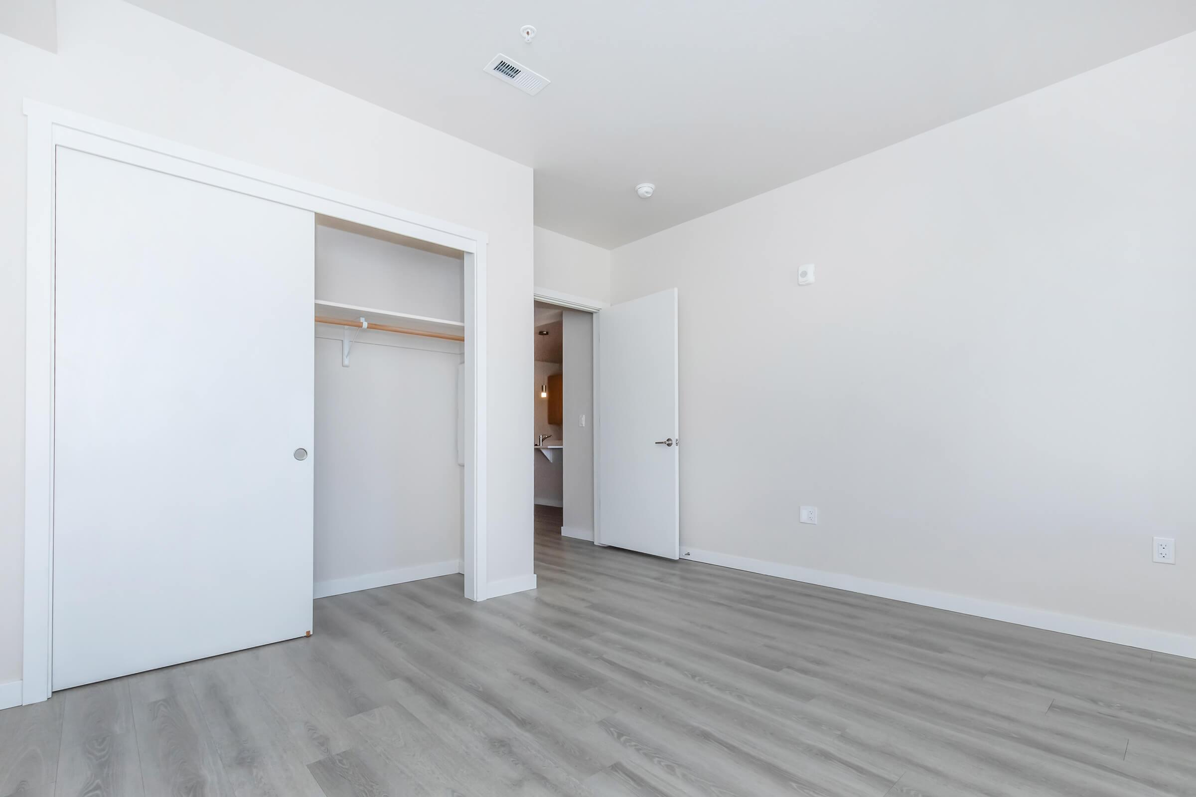 An empty room featuring light-colored walls and laminate flooring. There are two doors: one leads to a closet with a shelf, and the other is an entry door. The space is well-lit and minimalistic, with a neutral color palette enhancing the spacious feel.