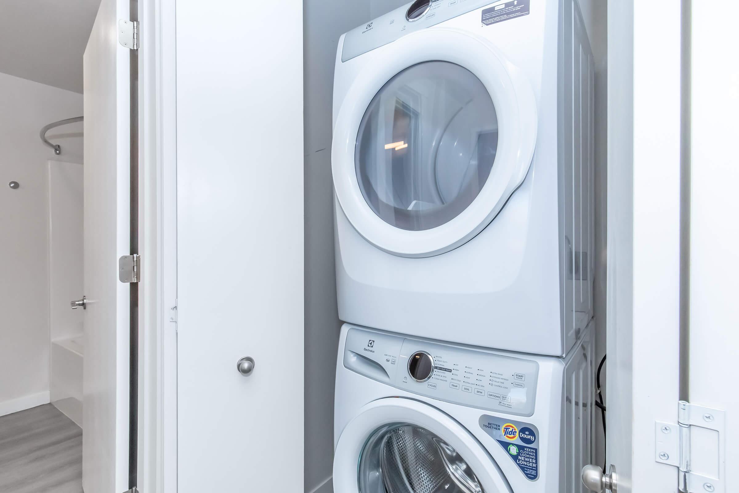 Stacked white washer and dryer located in a laundry closet. The appliances are front-loading, with a sleek design and control panel visible on the front of the washer. The space is well-lit and features a light-colored floor, with a white door partially closed on the left side, suggesting a compact laundry area in a home.