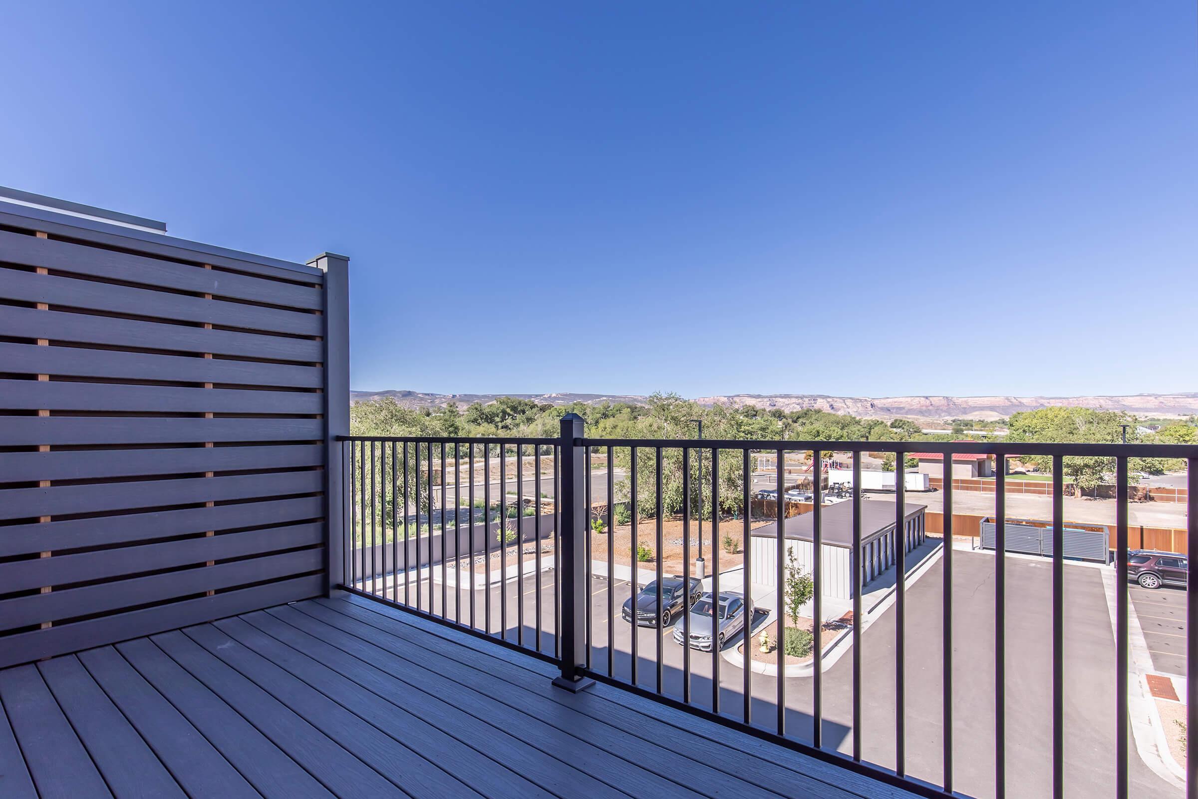 View from a balcony featuring a wooden railing, overlooking a parking area and landscape with distant mountains. The sky is clear and blue, creating a bright and open atmosphere.