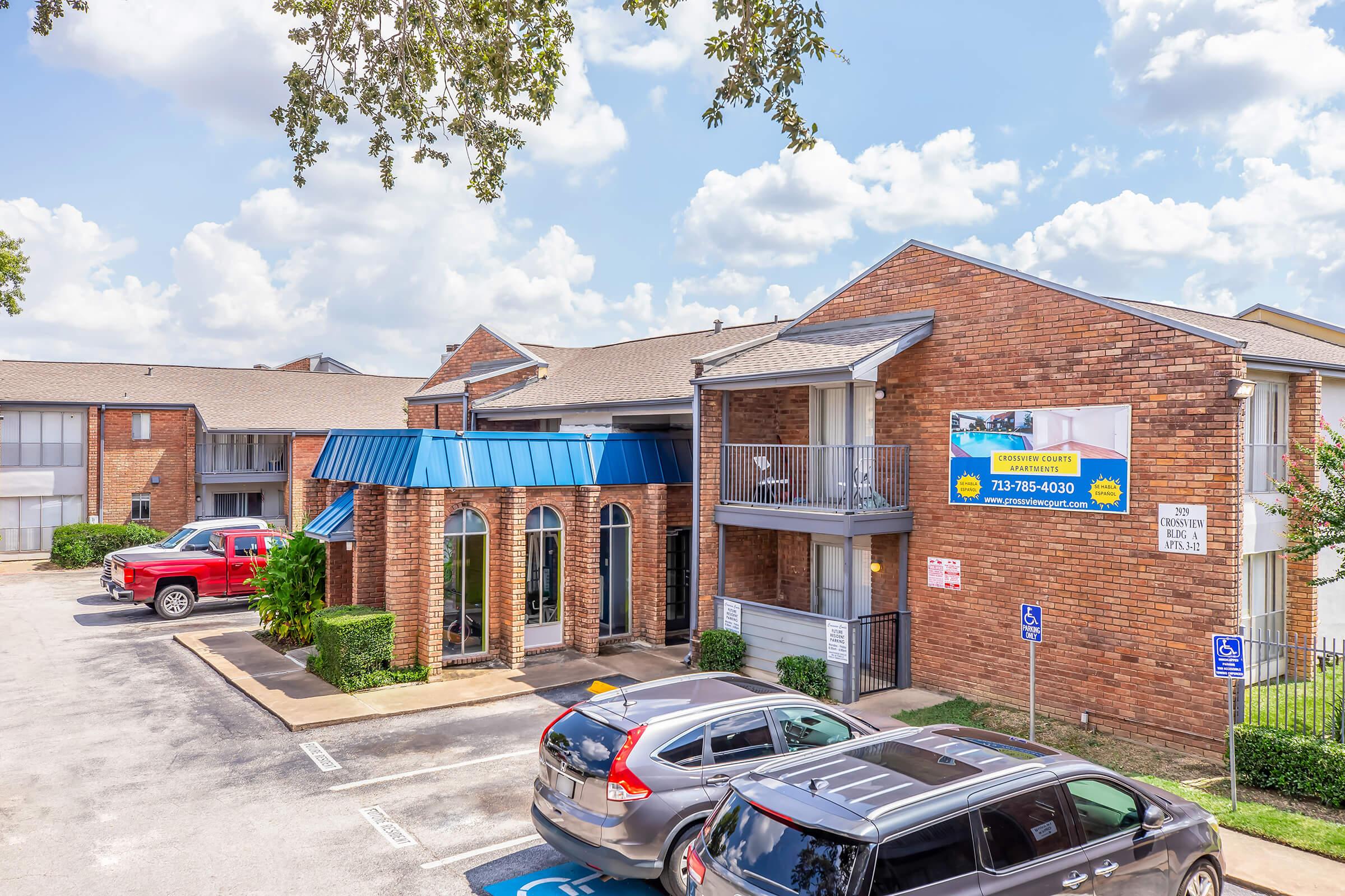 A brick apartment building with a blue peaked roof and a sign, surrounded by parked cars in a well-maintained lot. The sky is partly cloudy, and there are trees nearby. The structure features balconies and manicured landscaping, indicating a residential area.