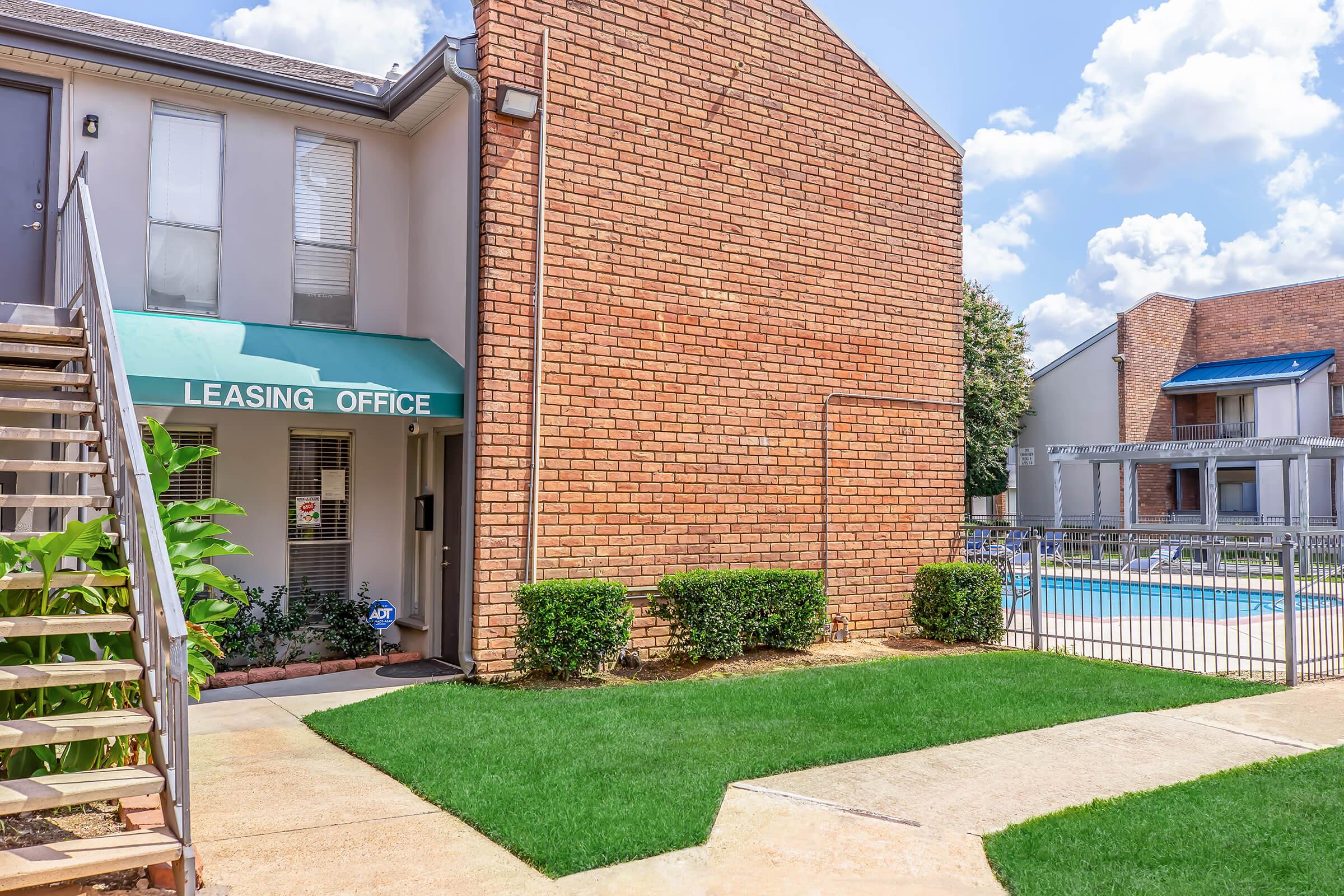 A leasing office entryway with a green awning, a staircase leading up on the left, and neatly trimmed bushes. In the background, a swimming pool is visible, framed by a fence. The sky is partly cloudy, creating a bright and inviting atmosphere.