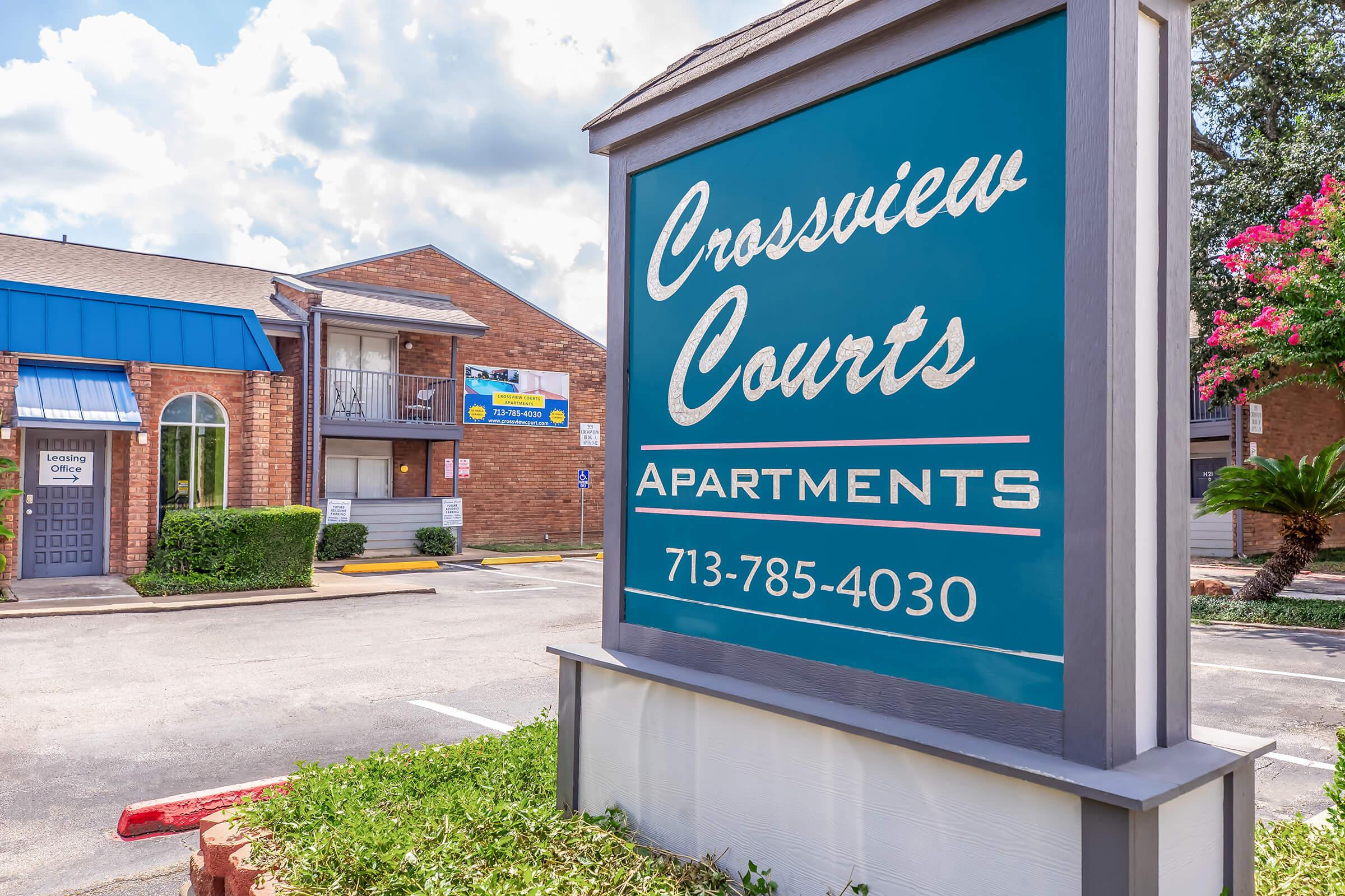 Sign for Crossview Courts Apartments displaying the name and contact number. The building features a mix of brick and wood siding, with a blue awning over the entrance. Surrounding landscaping includes palm trees and colorful flowers, set against a partly cloudy sky.