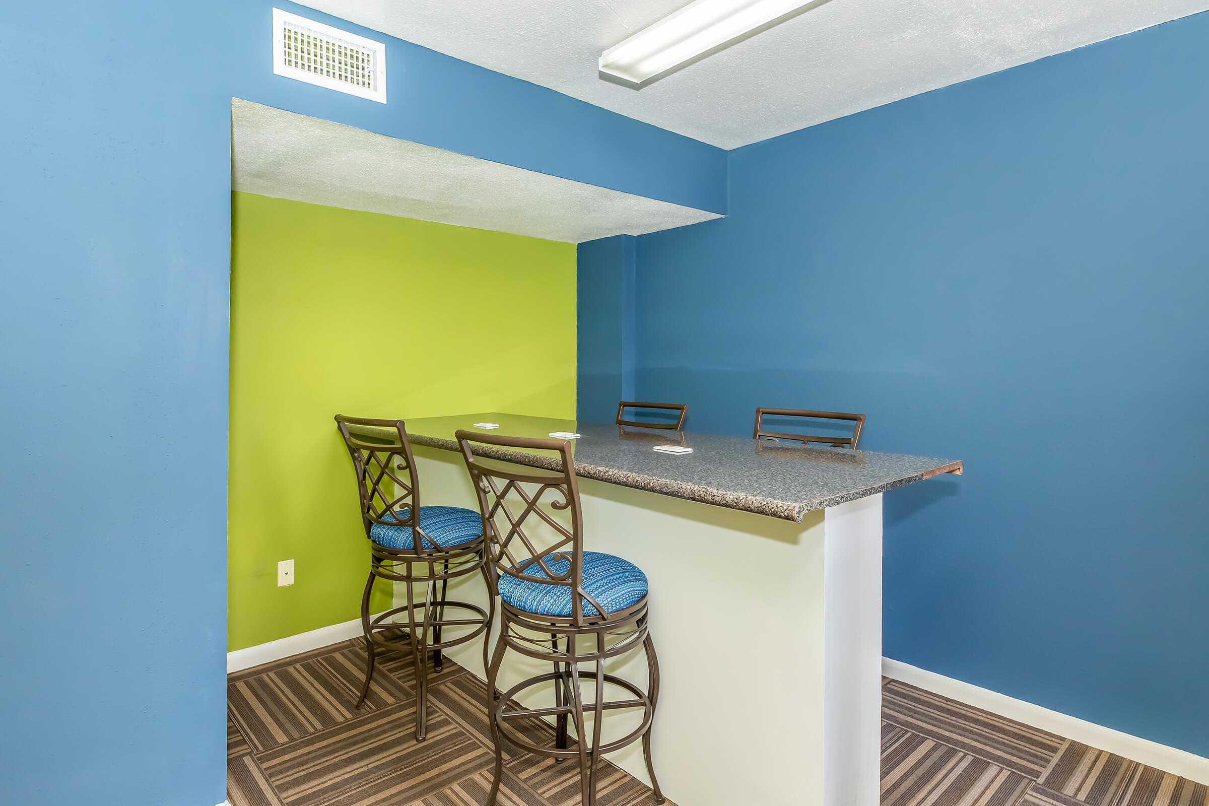 A modern kitchen bar area featuring a granite countertop, two metal bar stools with blue upholstery, and colorful walls painted in blue and lime green. The flooring consists of striped carpet, creating a stylish and inviting atmosphere.