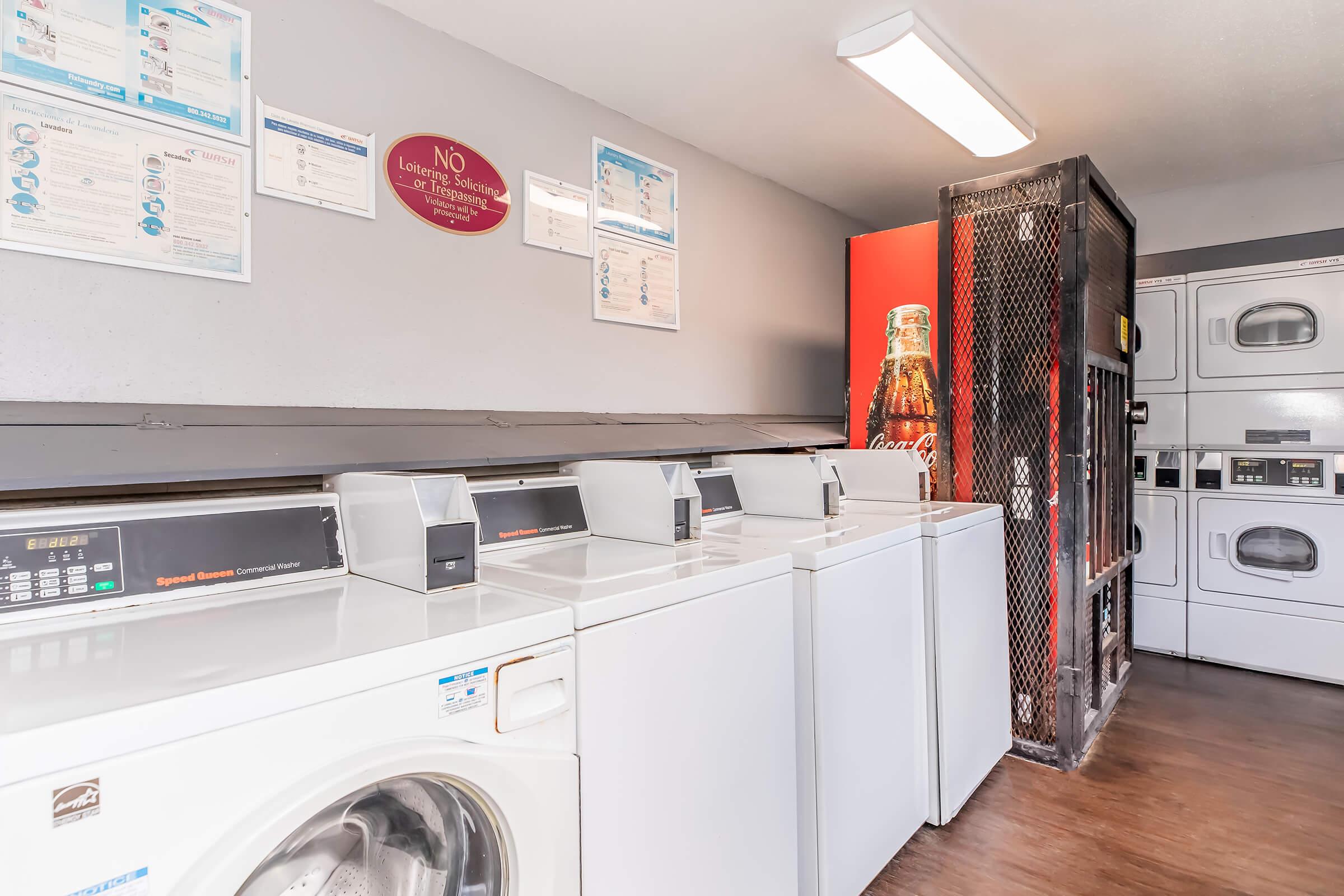 A laundry room featuring several white washing machines and dryers arranged in a row. Above the machines, there are informational posters and a wall-mounted sign stating "No Laundry." On one side, there's a Coca-Cola vending machine. The floor is wooden and well-maintained.