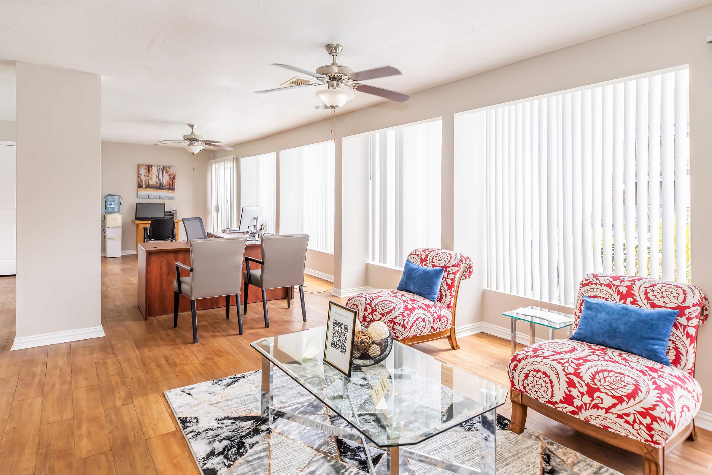 Bright and airy living space featuring two patterned armchairs, a glass coffee table with a decorative item, and a desk area with modern office chairs. Large windows allow natural light to fill the room, enhancing the open and inviting atmosphere. The flooring is a warm wood laminate.