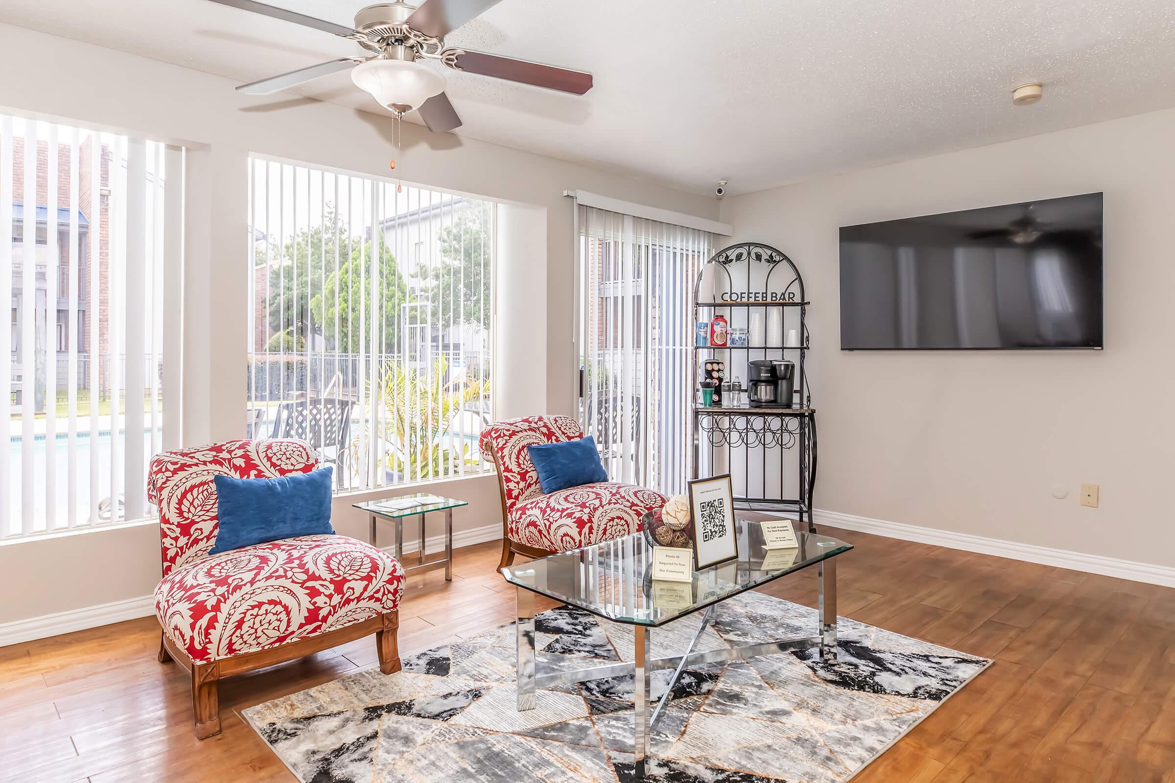 Bright and inviting living room featuring two red patterned chairs with blue cushions, a glass coffee table on a patterned rug, and a black metal bar cart. Large windows with blinds let in natural light, and a wall-mounted TV is visible. The decor is modern and comfortable, creating a cozy atmosphere.