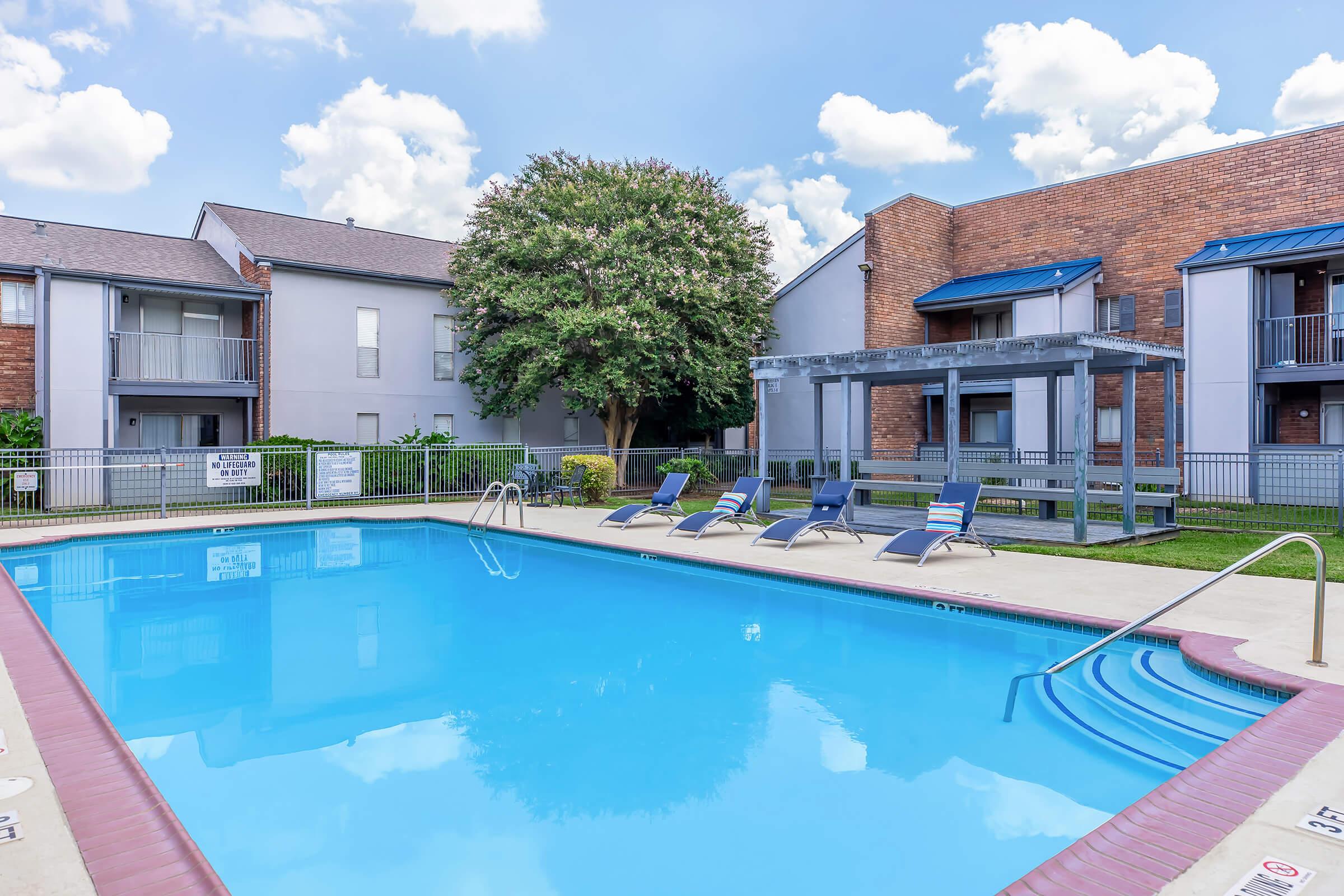 A clear blue swimming pool surrounded by lounge chairs, with a shaded gazebo nearby. The pool area is located in a residential complex, featuring several brick and modern buildings in the background under a partly cloudy sky. Green shrubs and trees add greenery to the scene.