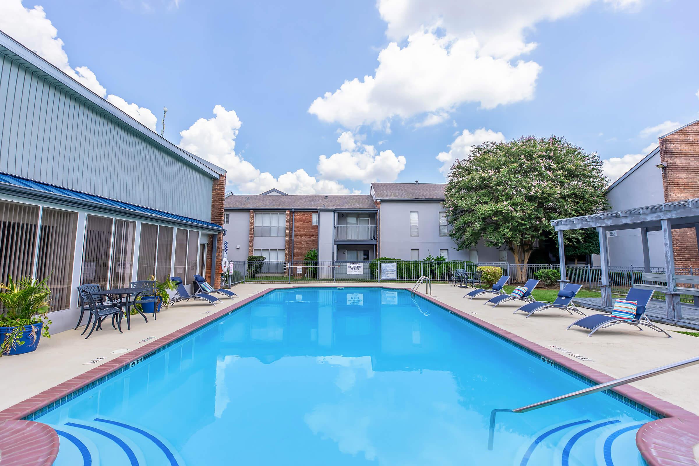 A clear blue swimming pool surrounded by lounge chairs and a shaded seating area. In the background, there are buildings with balconies and green landscaping under a partly cloudy sky.