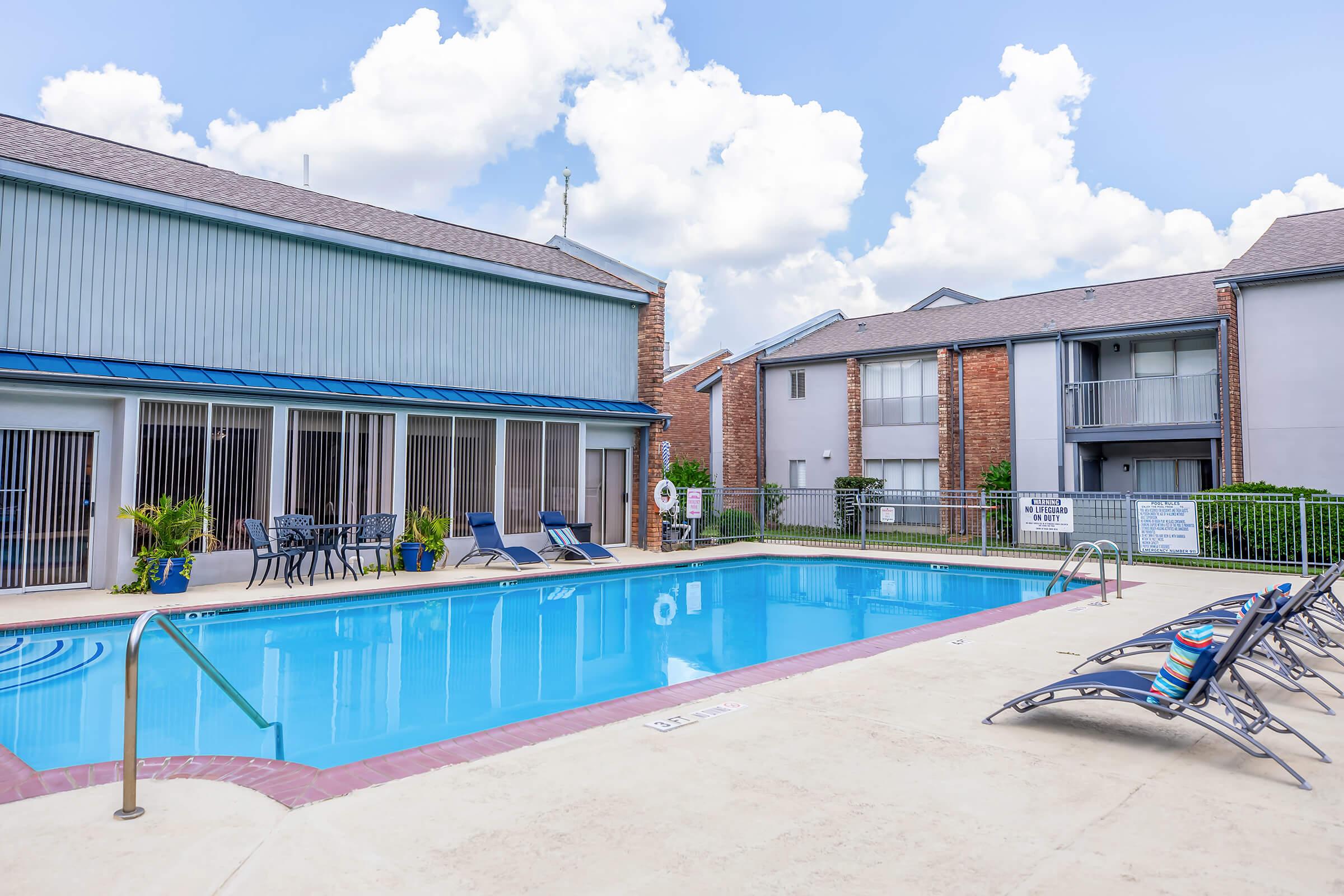 A well-maintained swimming pool area featuring blue water, lounge chairs, and an inviting atmosphere. Surrounding structures include a modern building with large windows and a brick facade, set against a backdrop of fluffy white clouds and a clear blue sky.