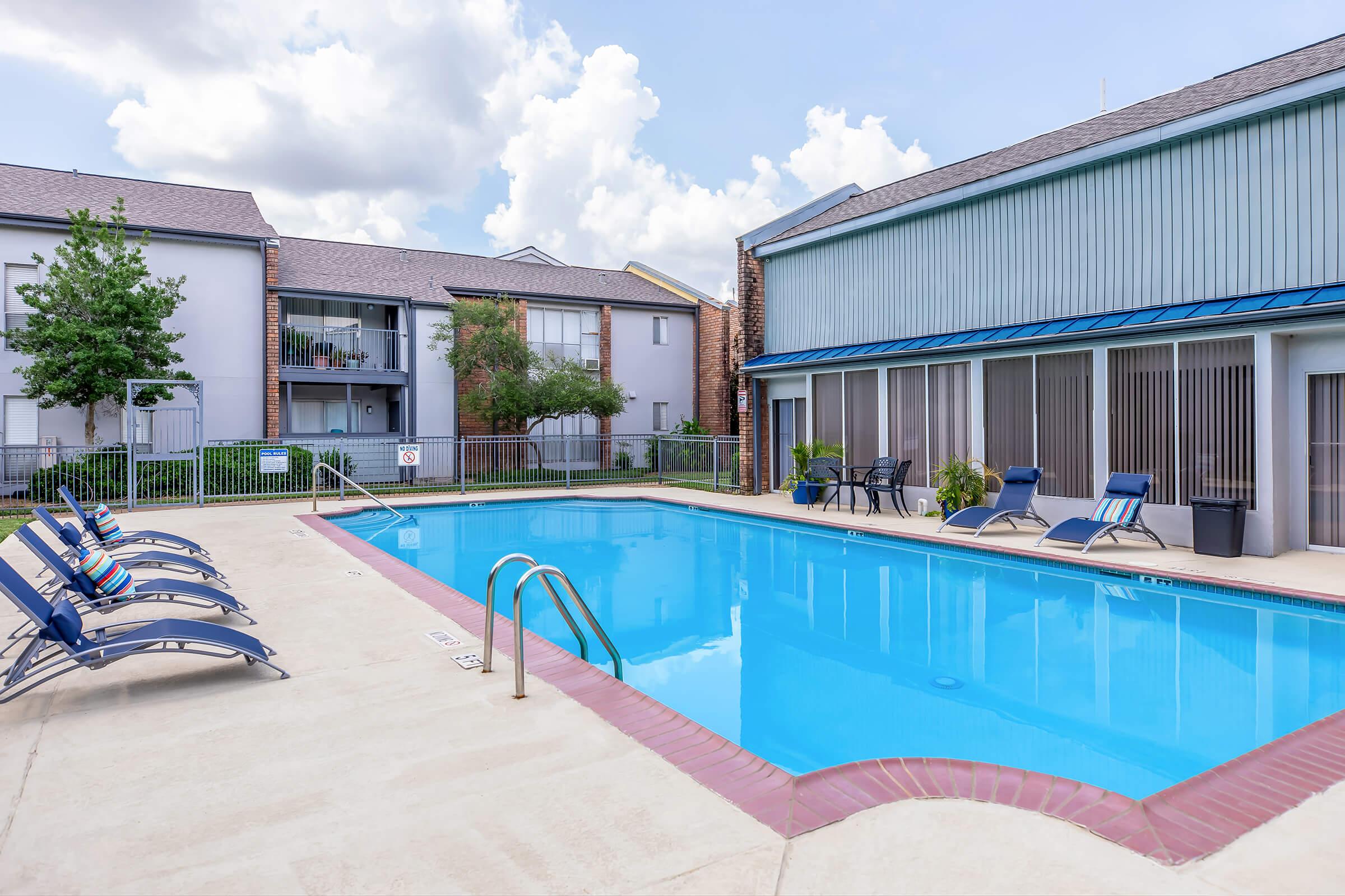 A clear blue swimming pool surrounded by lounge chairs, with residential buildings in the background. The pool area is well-maintained, featuring a fence and greenery. The sky is partly cloudy, creating a bright and inviting atmosphere.