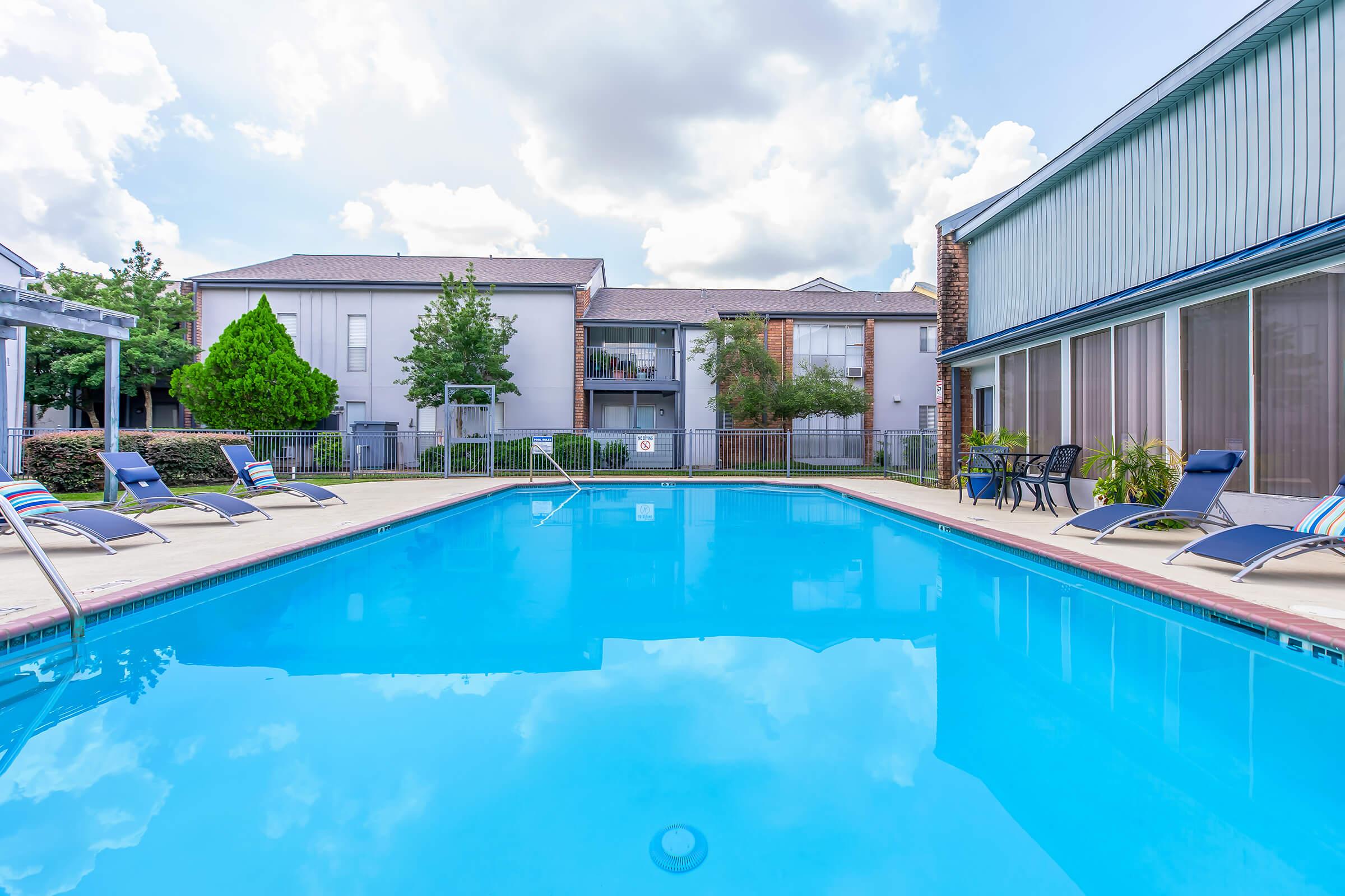 A clear blue swimming pool surrounded by lounge chairs, with apartment buildings in the background. Lush trees and green landscaping enhance the setting, while fluffy clouds partially cover a bright sky. The scene conveys a peaceful and inviting atmosphere for relaxation.