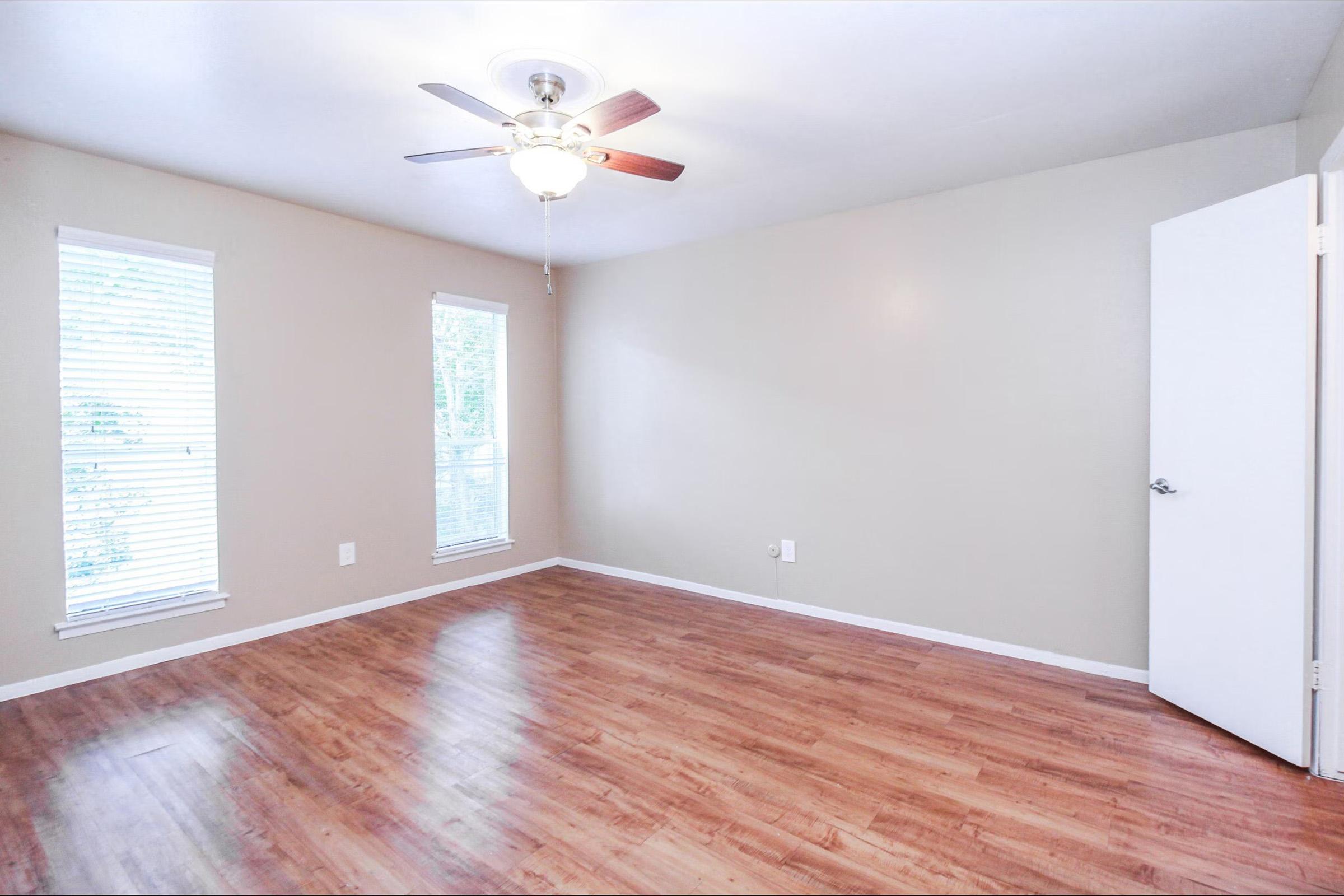A spacious, empty room featuring light brown walls and wood-style flooring. There are two windows with blinds allowing natural light, and a ceiling fan is mounted in the center. A white door on the right leads to another space, creating a bright and airy atmosphere.