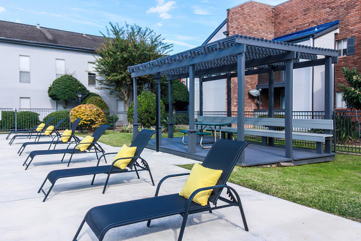 A sunny outdoor pool area featuring several lounge chairs with yellow cushions, a shaded gazebo structure, and landscaped greenery in the background, alongside a modern building.