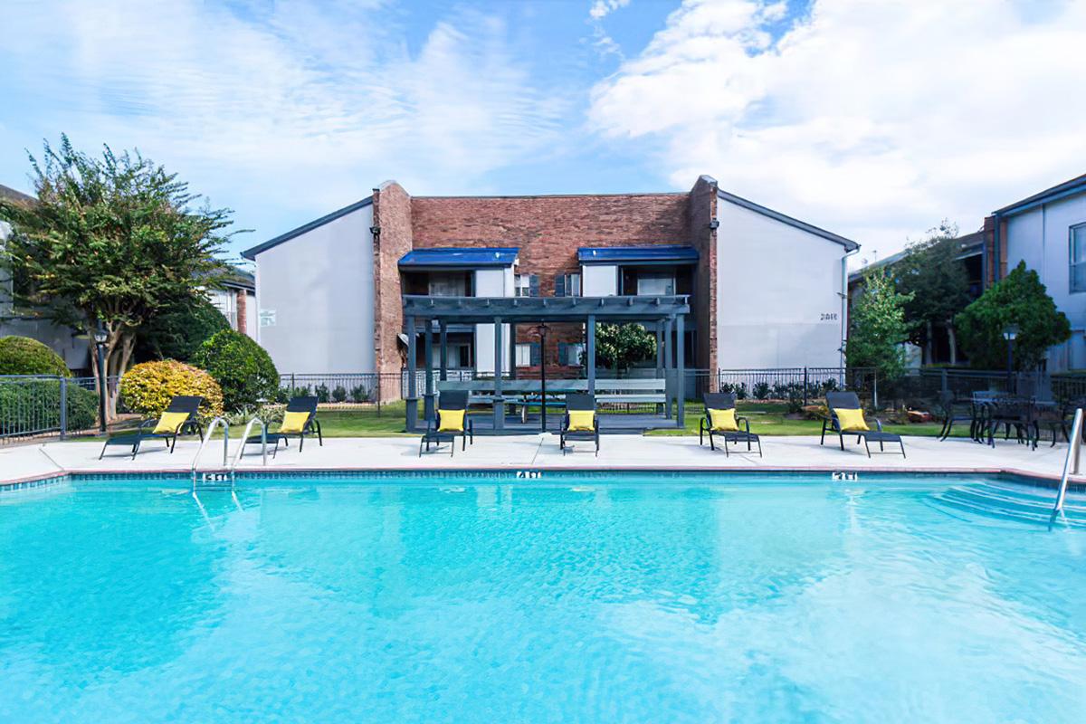 A swimming pool surrounded by lounge chairs and greenery, with a modern apartment building in the background. The sky is partly cloudy, creating a bright and inviting atmosphere. The pool area is well-maintained, offering a relaxing space for residents.