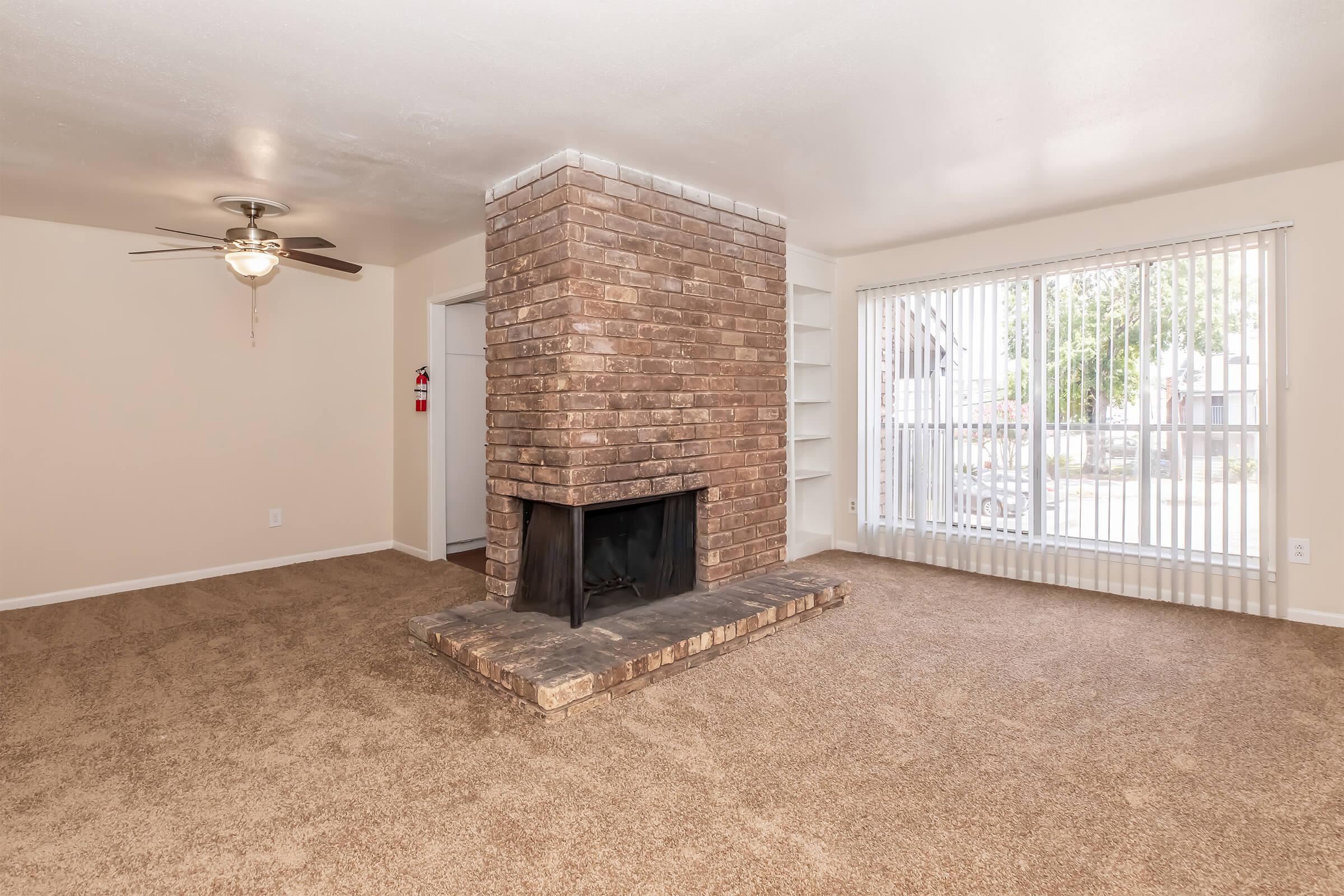 Spacious living room featuring a brick fireplace as the focal point, beige walls, and plush brown carpet. A ceiling fan provides ventilation, while large windows allow natural light to fill the space. Built-in shelving is visible, adding to the room's functionality and charm.