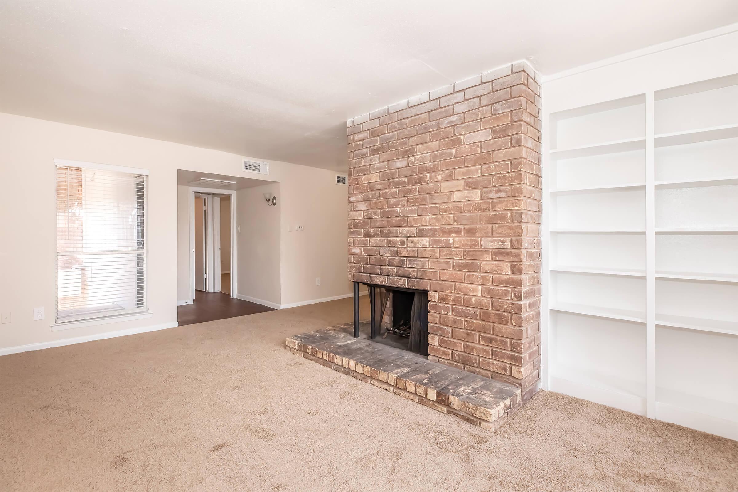 A spacious living room featuring a brick fireplace with a stone hearth, beige carpet, and light-colored walls. There's a built-in bookshelf to the right and a doorway leading to another room. Natural light flows through a window, creating a bright and inviting atmosphere.