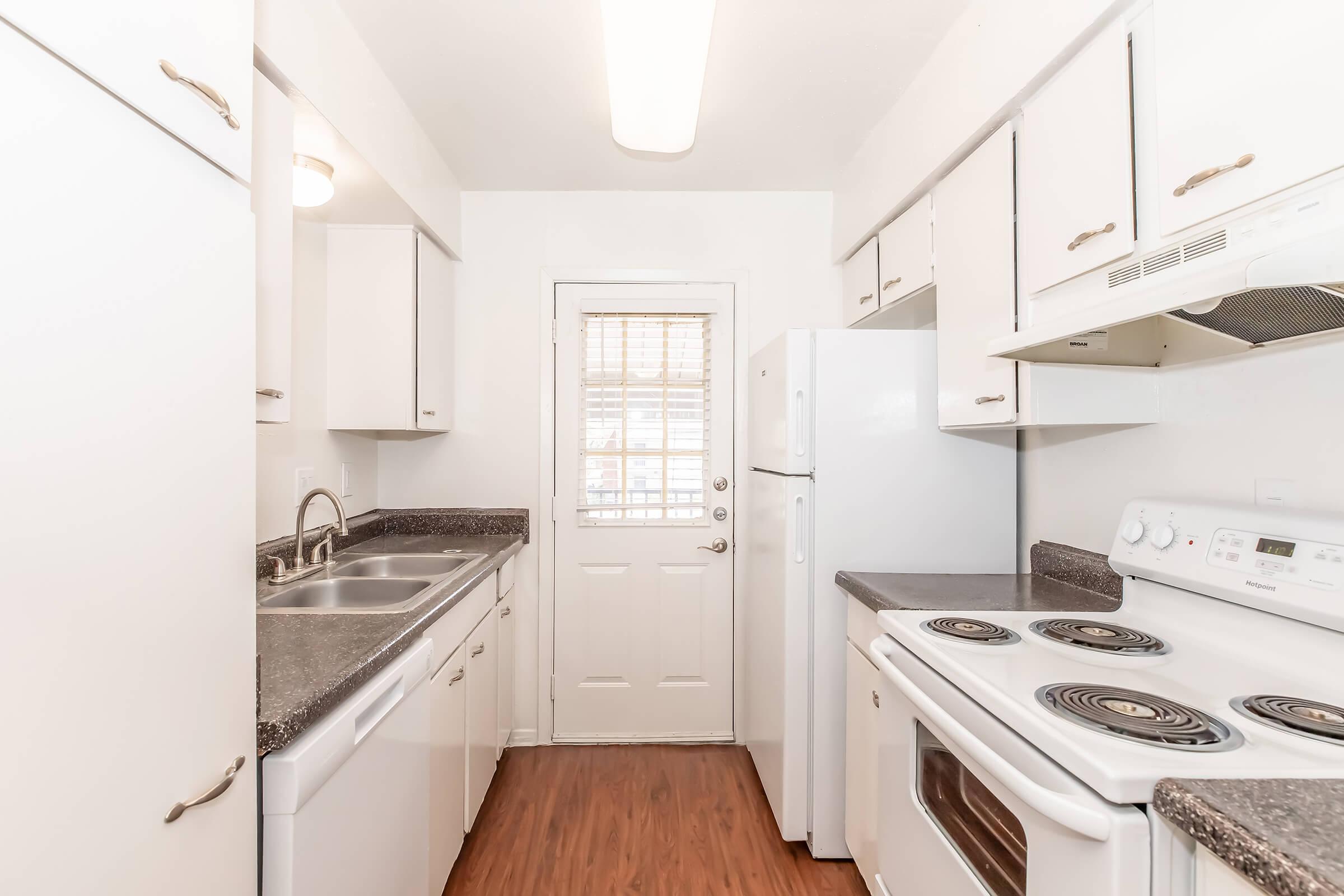 A modern kitchen with white cabinetry, a double sink, and stainless steel appliances including a dishwasher and stove. The flooring is wood-like, and there's a door leading outside. Natural light enters through a window with blinds, creating a bright and tidy atmosphere.