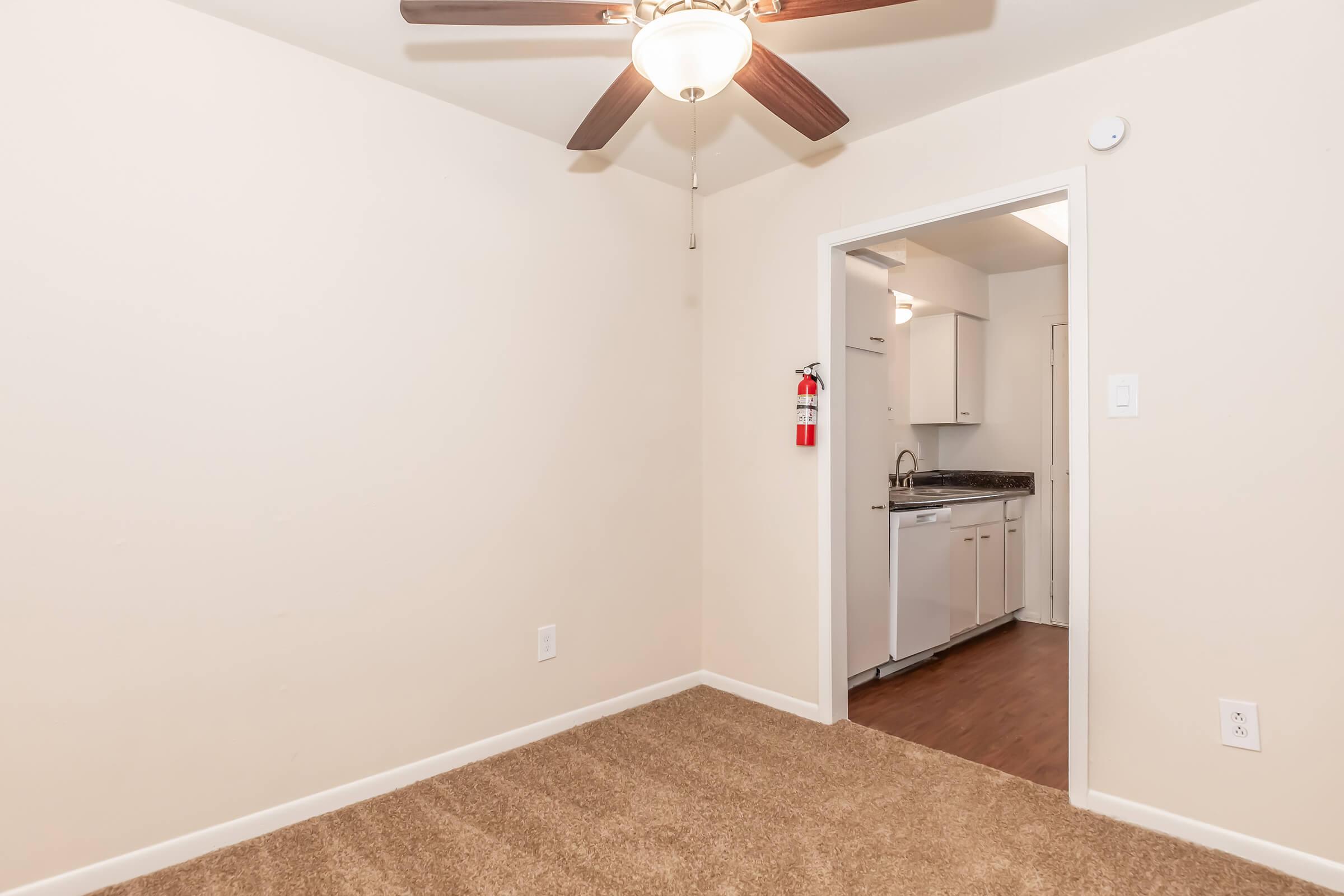 A small empty room with light beige walls and a ceiling fan. The floor is carpeted in a light brown shade. A doorway leads to a kitchen area featuring white cabinets and appliances. A fire extinguisher is mounted on the wall near the entrance to the kitchen.