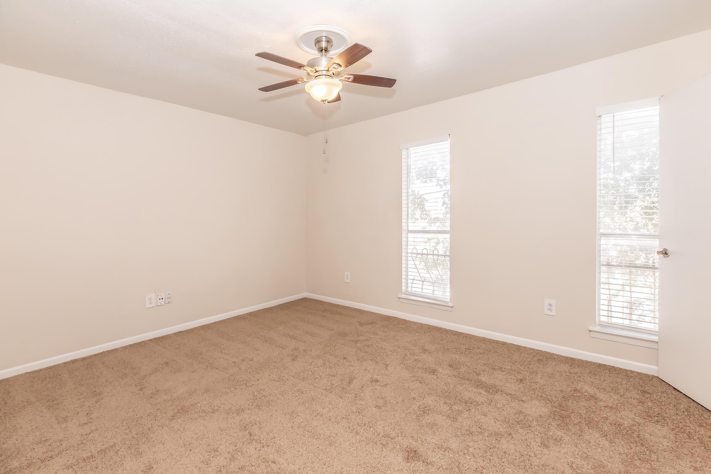 A spacious, empty room with beige walls and carpet. It features a ceiling fan with light, two windows allowing natural light in, and a white door. The room is well-lit and has a clean, minimalist appearance.