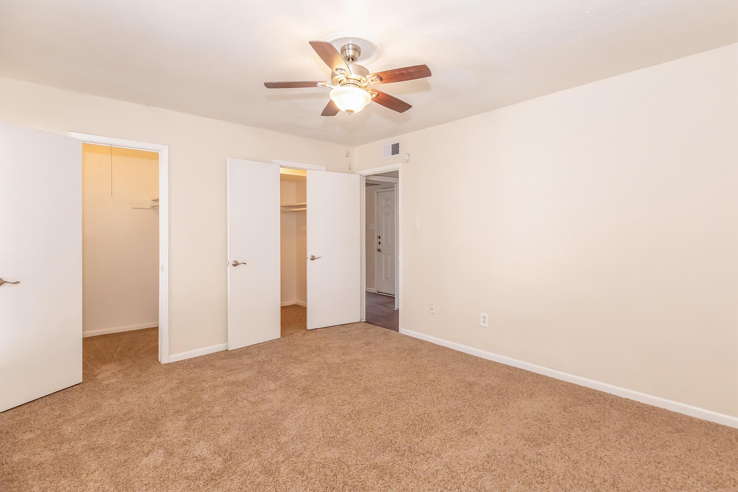 A well-lit room featuring beige walls and carpet flooring. There is a ceiling fan with light, two white closet doors, and a doorway leading to another space. The room is empty, creating a spacious and inviting atmosphere.