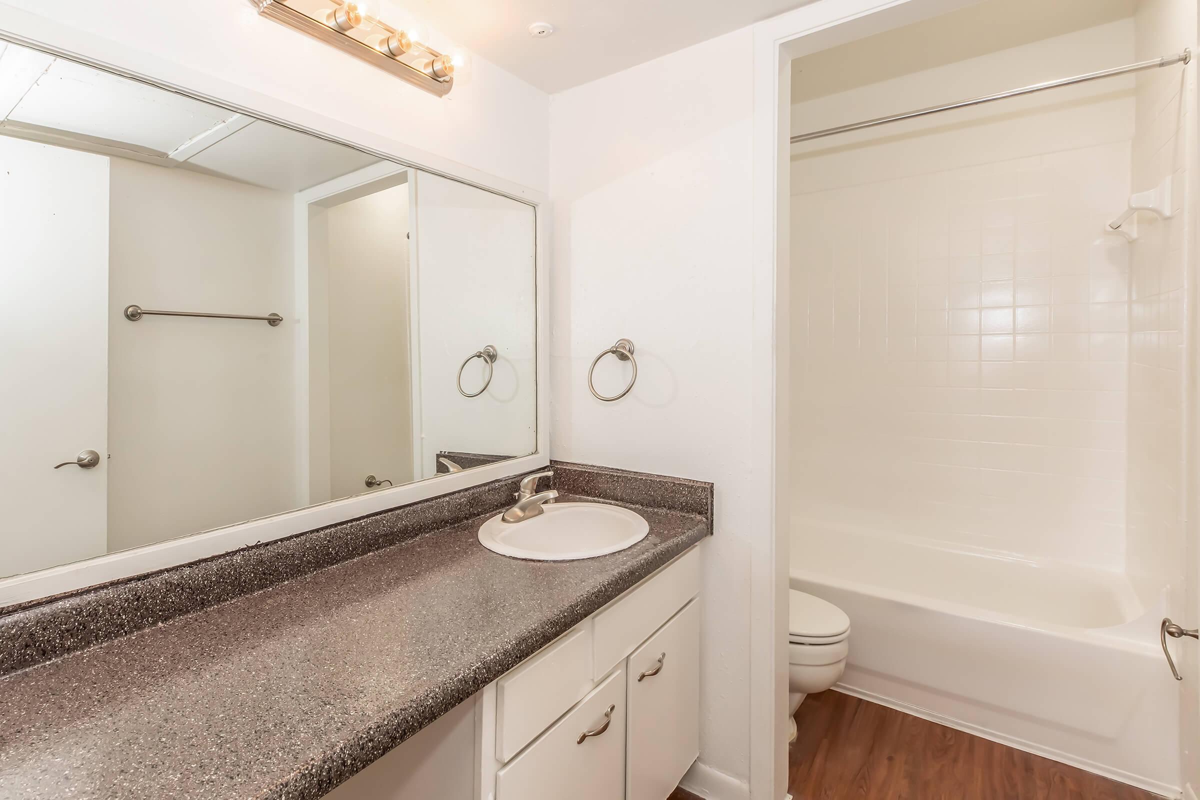 A clean, well-lit bathroom featuring a double sink vanity with a dark countertop, a large mirror above the sink, and a shower/tub combo. The walls are painted white, and there is a towel bar on the wall. The flooring appears to be a wood-like laminate.