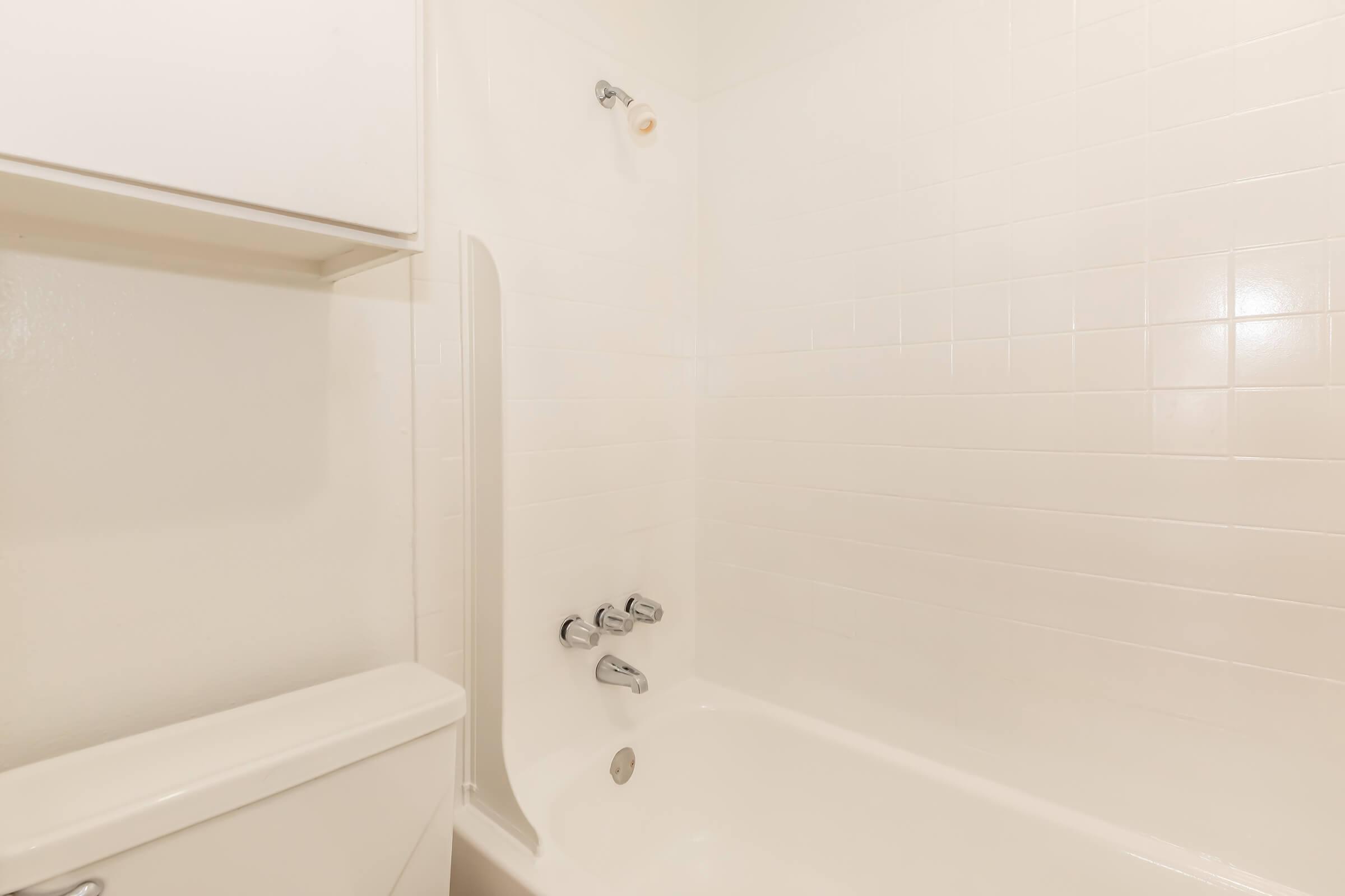 A clean, minimalist bathroom featuring a white bathtub, showerhead, and modern faucet. The walls are tiled in a light color, and there is a small shelf above the bathtub. A toilet is positioned next to the bathtub, contributing to the simple design of the space.