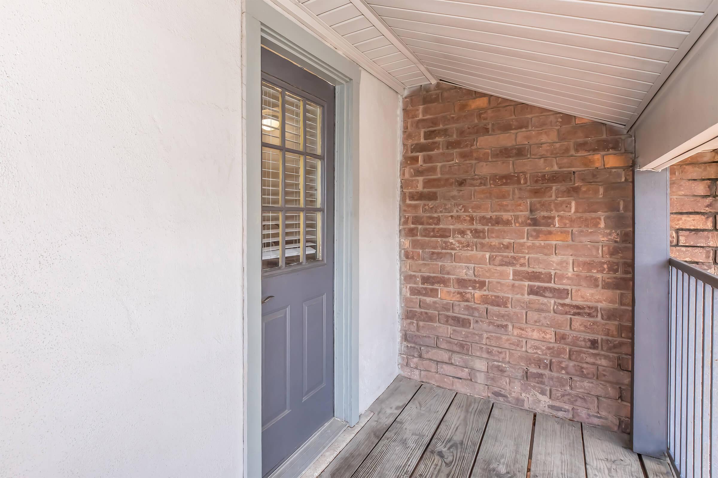 A small covered porch featuring a gray door with a window and wooden floorboards. The wall on one side is made of light-colored material, while the other side showcases exposed brick. The ceiling is white with a simple design, providing a cozy entrance to a home.
