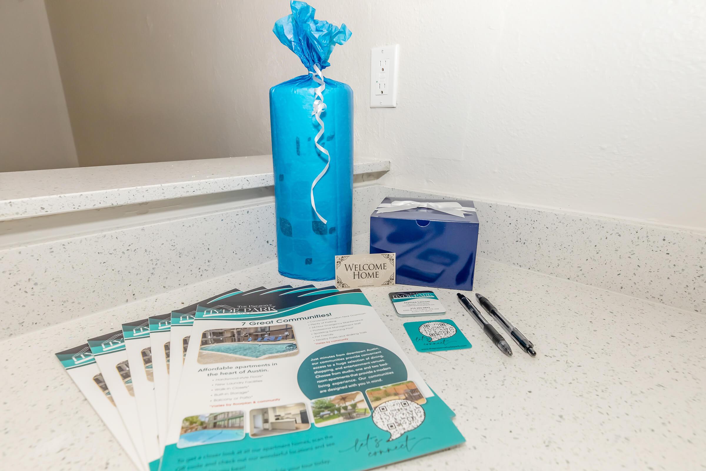 A neatly arranged welcome display on a countertop, featuring a blue-wrapped gift, brochures about a community, a "Welcome Home" sign, a blue box, and two pens. The scene conveys a warm, inviting atmosphere, likely for new residents or guests.