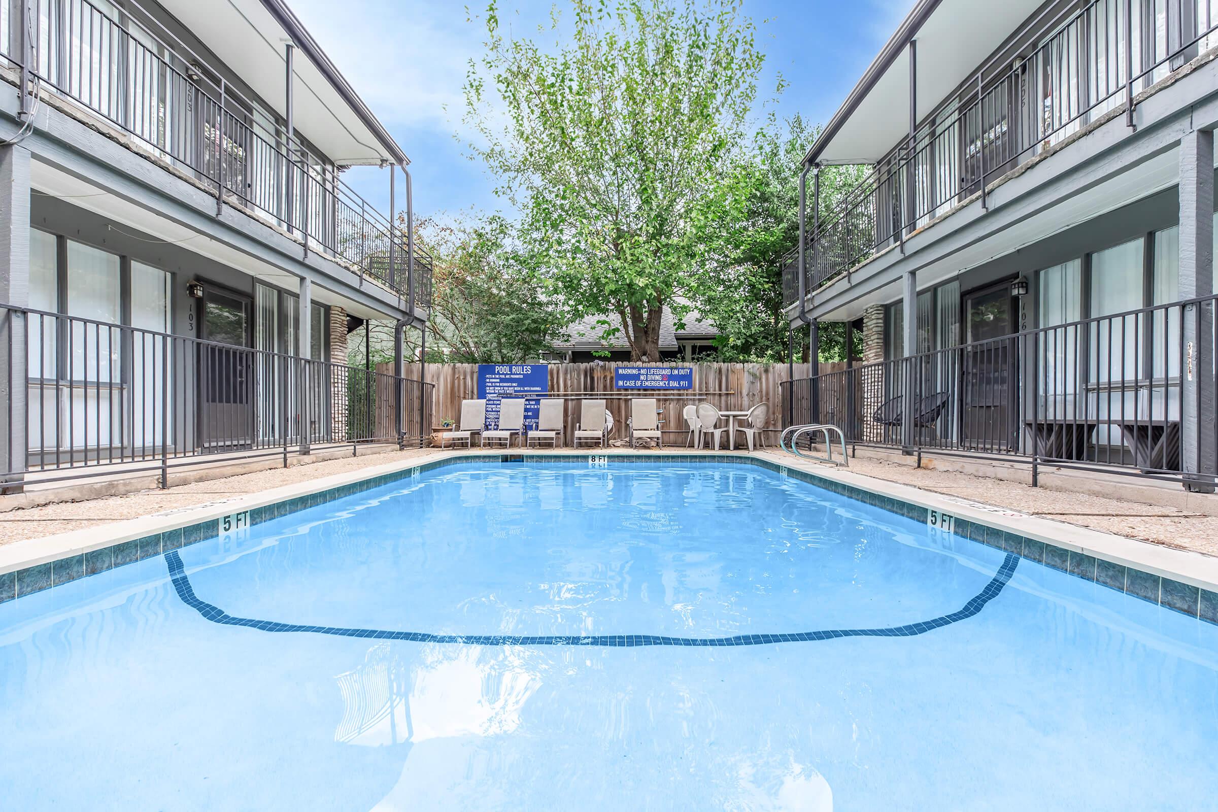 A clear, calm swimming pool surrounded by two-story apartment buildings. The pool area features lounge chairs and a safety sign. Lush greenery and trees are visible in the background, with a bright, blue sky overhead. The scene conveys a relaxing residential atmosphere.