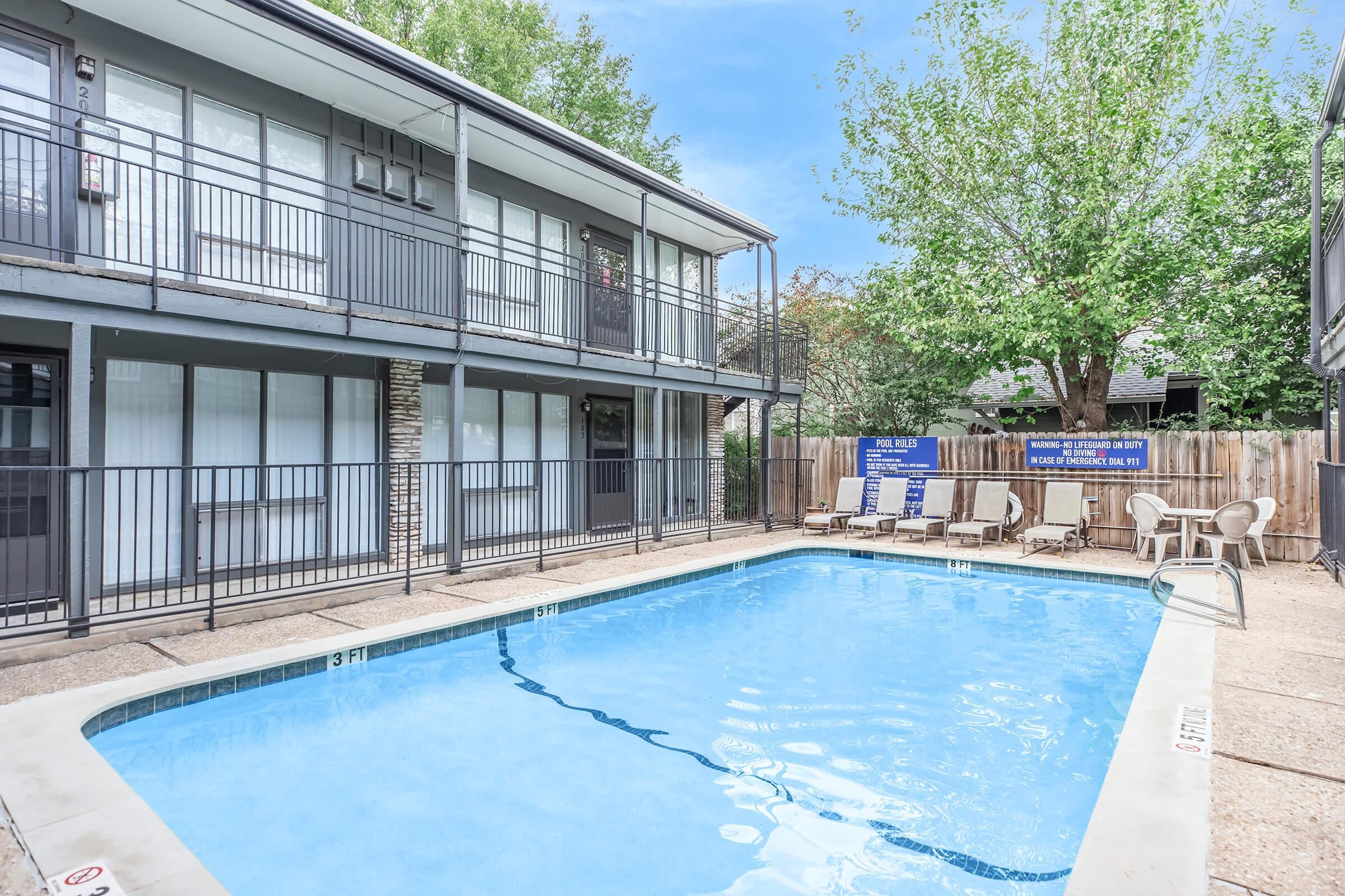 A clear blue swimming pool surrounded by a patio area, with lounge chairs and signs indicating rules. The pool is situated next to a multi-story building with balconies. Lush green trees can be seen in the background, enhancing the relaxing atmosphere of the space.