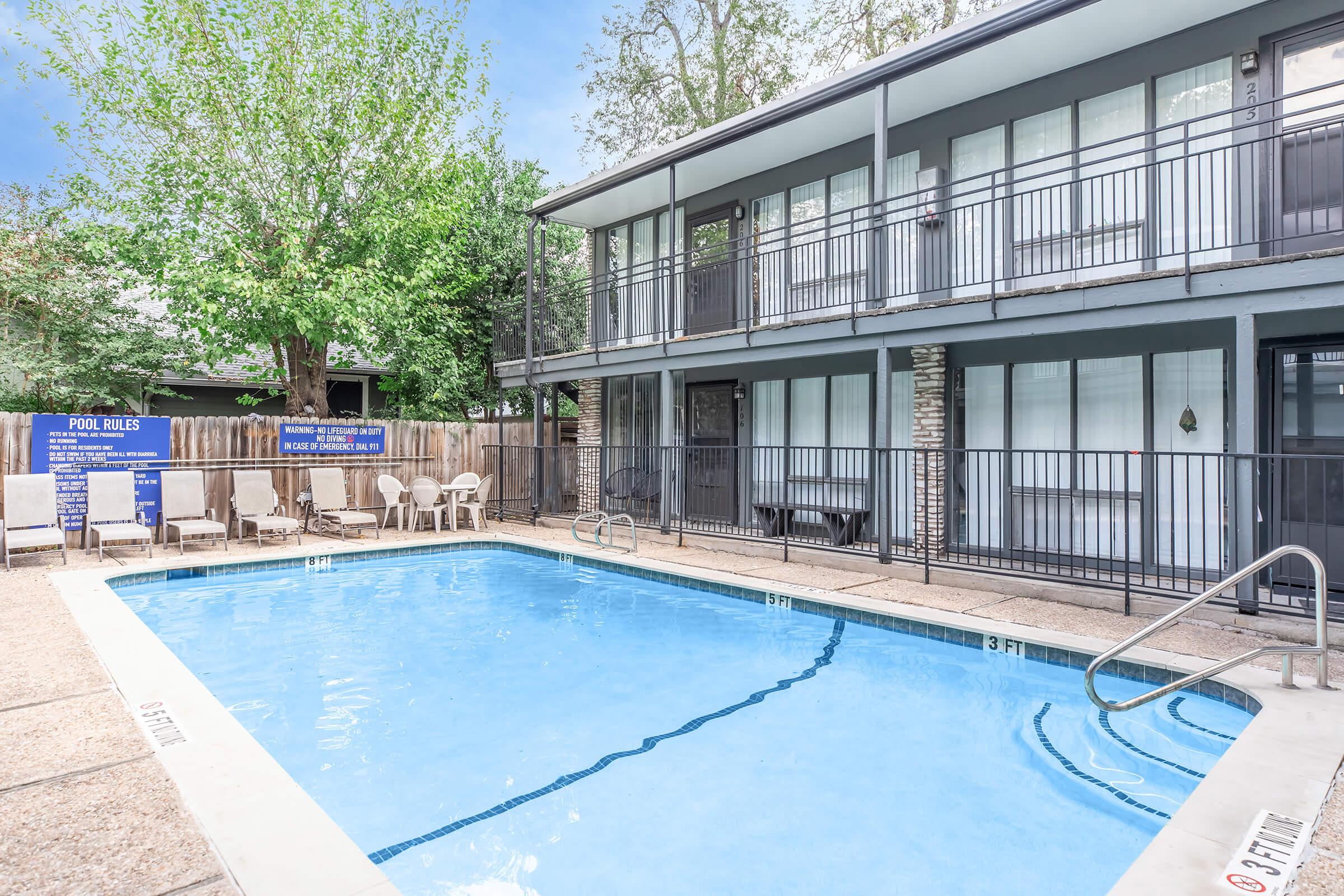 A clear, inviting swimming pool surrounded by a stone patio and chairs, with a two-story building in the background. The area is shaded by trees, and there's a sign visible with pool rules. The scene is bright and welcoming, ideal for relaxation and leisure.