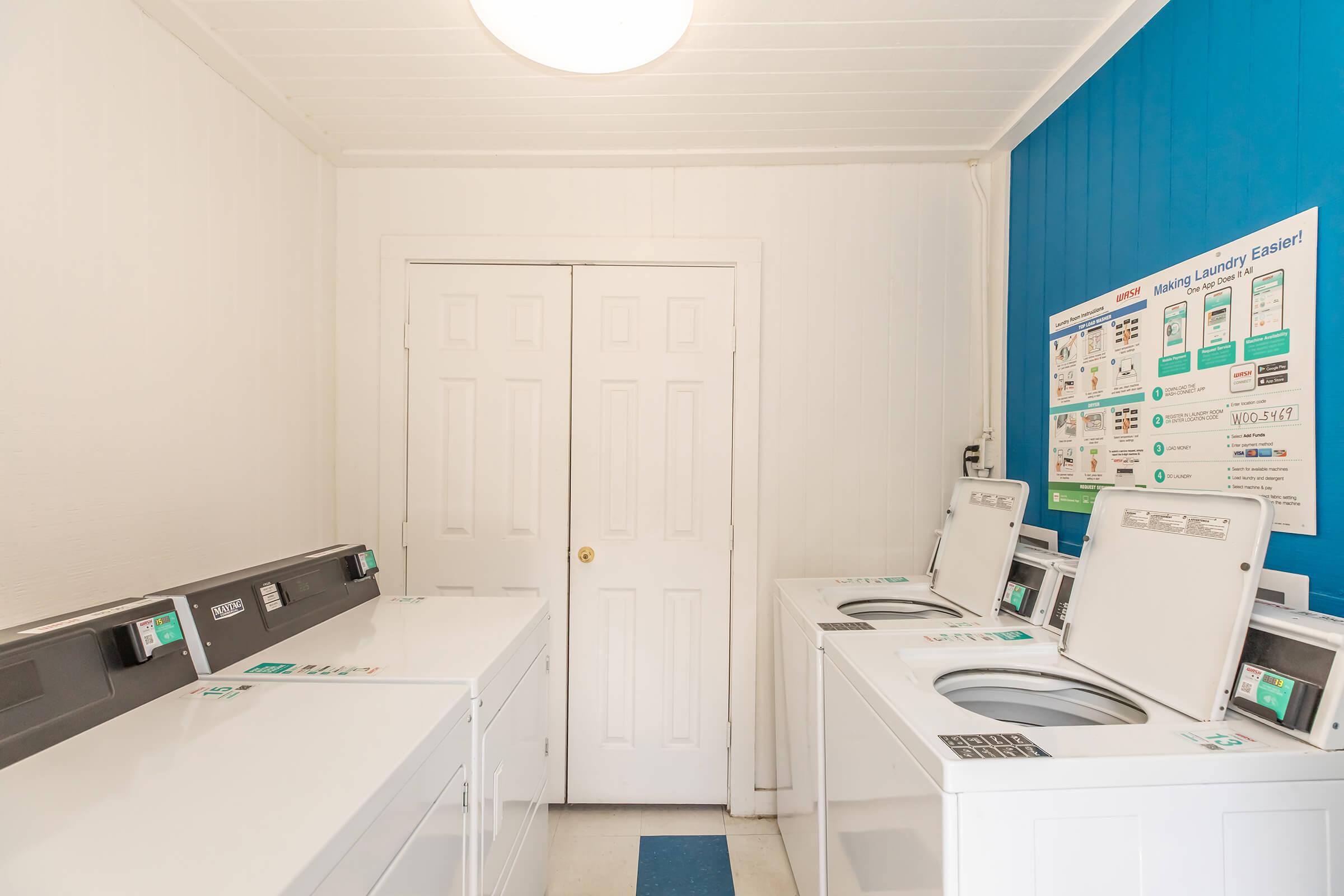 A clean and modern laundry room featuring multiple white washers and dryers arranged against a light-colored wall with a blue accent. A door leads to another room, and there are informational posters on the wall detailing laundry services. The floor is tiled with a blue and white pattern.