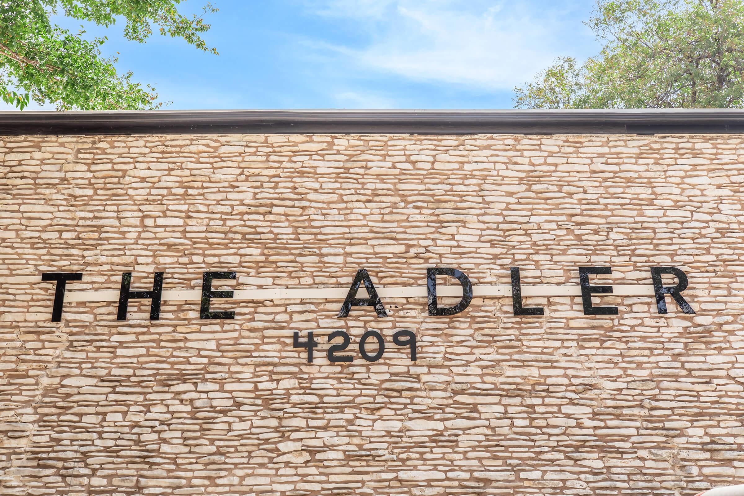 A stone wall featuring the text "THE ADLER" in large, black letters, along with the number "4209." The background includes a clear blue sky and green foliage from trees, adding a natural touch to the urban setting.