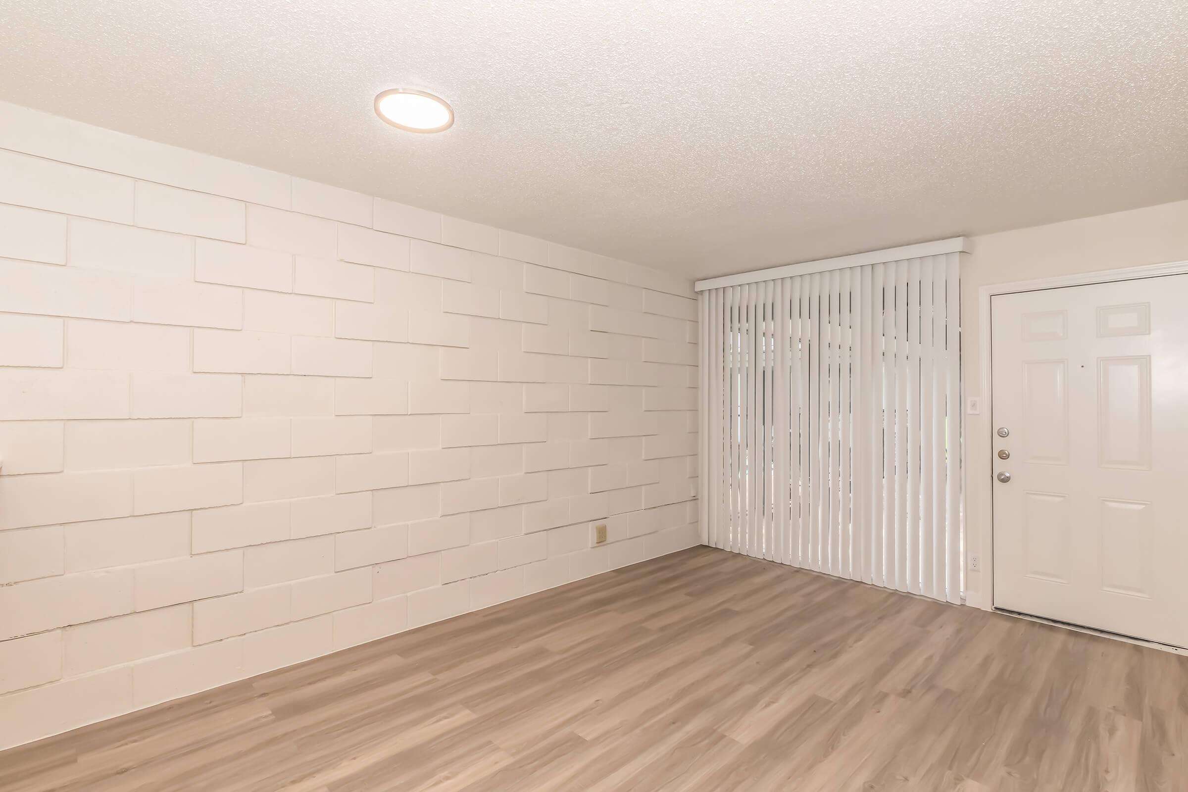 An empty room with light-colored walls featuring a textured design. The floor is covered in light wood laminate. There is a sliding glass door with vertical blinds on one side and a simple white door with a doorknob on the other. The ceiling is equipped with a round light fixture.