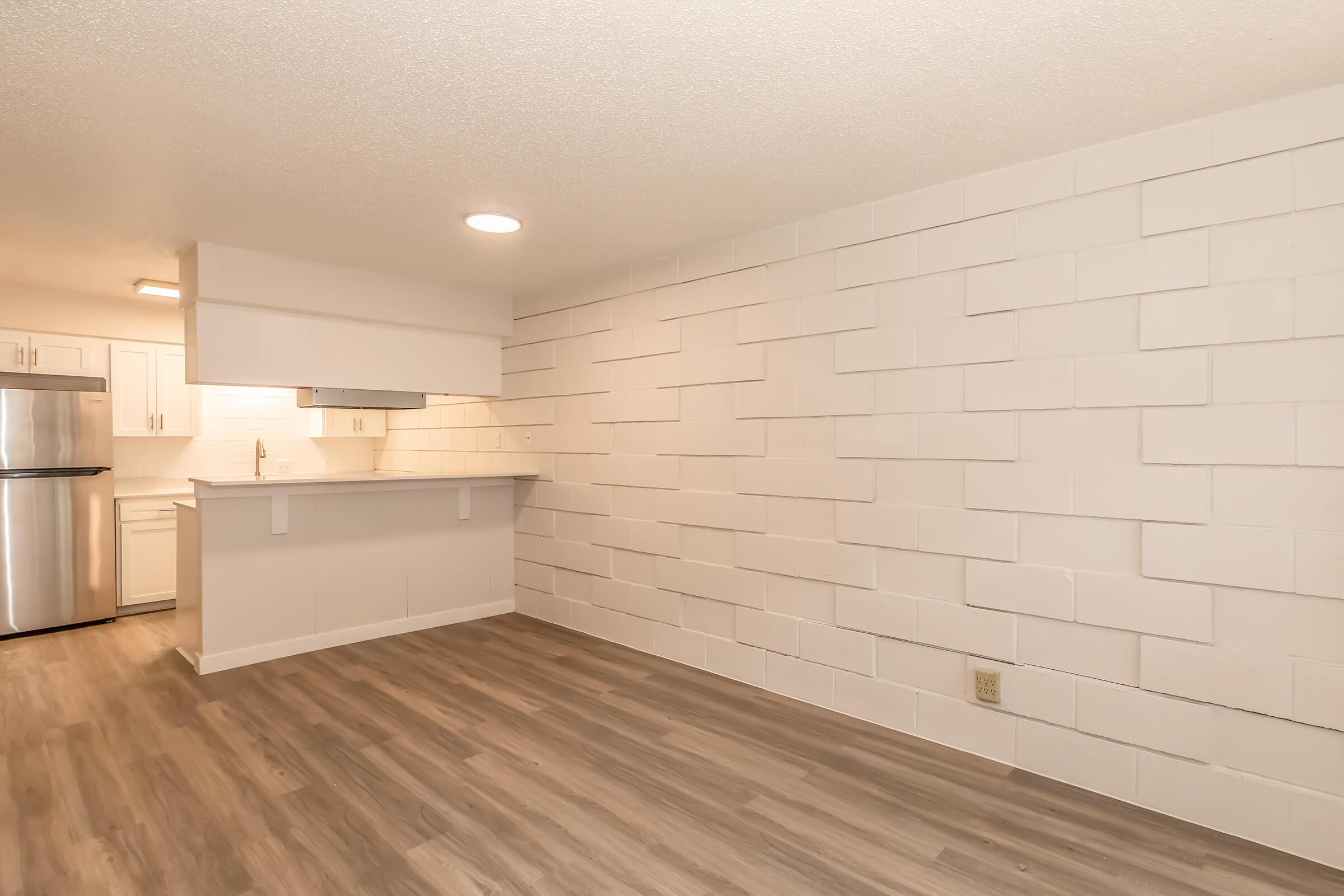 A modern, unfurnished living space featuring a light-colored tiled wall and new flooring. The open area leads to a kitchen with white cabinetry and stainless steel appliances, illuminated by ceiling lights. The overall ambiance is bright and airy, suitable for various interior design options.
