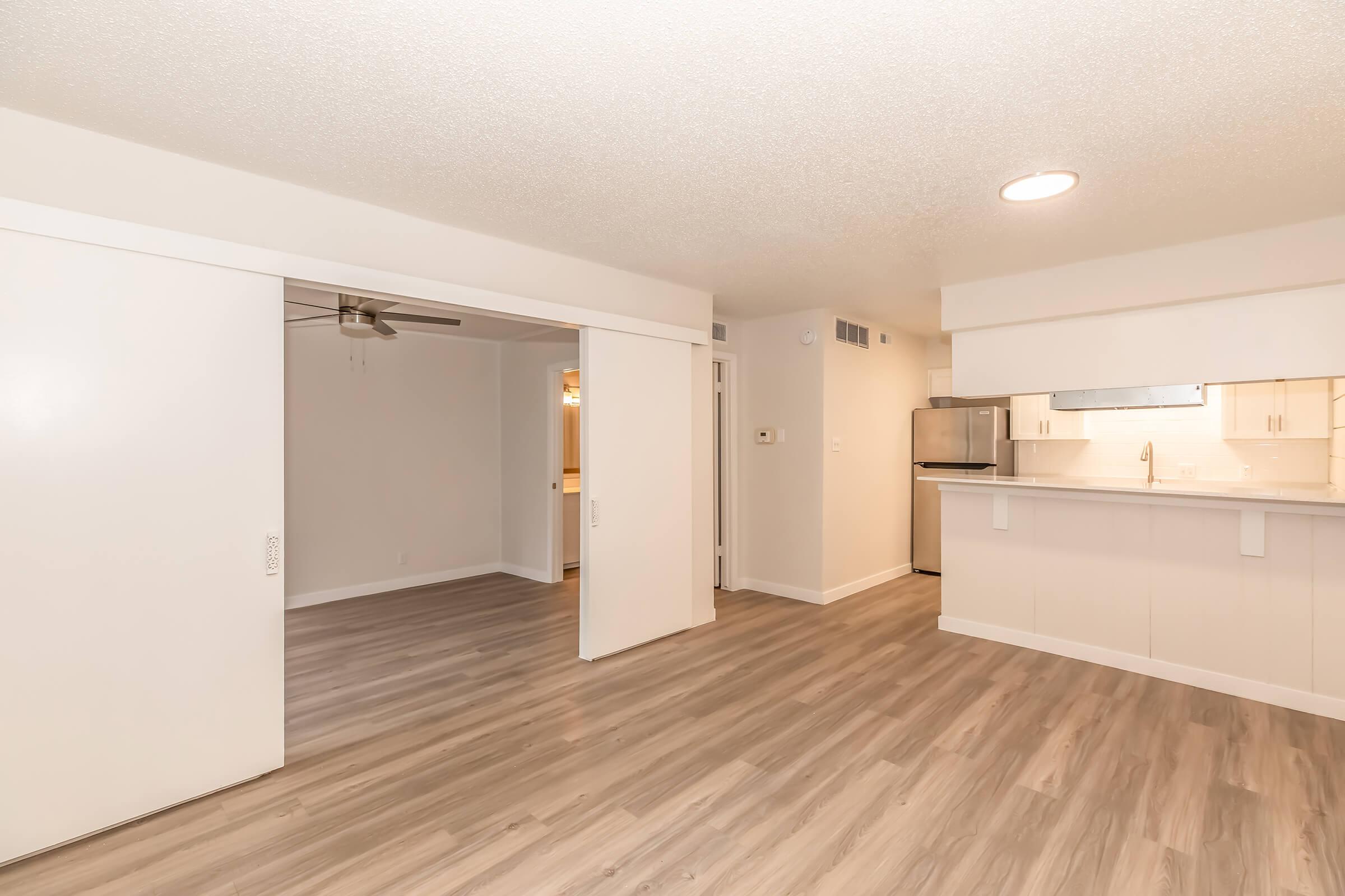 Interior view of a modern apartment showcasing a spacious living area with light-colored walls, laminate flooring, and a ceiling fan. A set of sliding doors leads to an adjacent room, and the kitchen is visible in the background, featuring white cabinetry and stainless steel appliances. Bright, airy atmosphere.