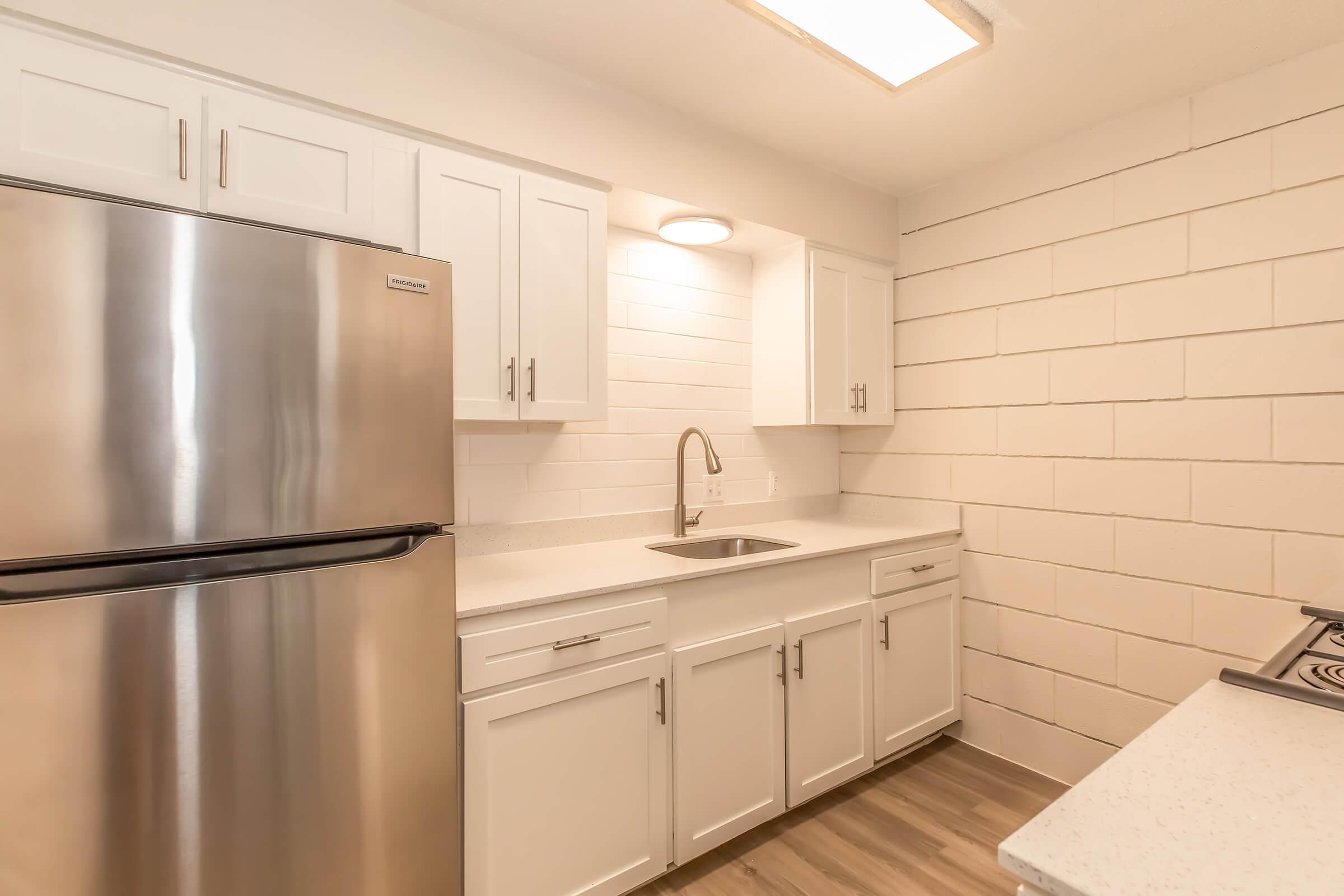 Modern kitchen with stainless steel refrigerator, white cabinets, and a countertop sink. The walls feature a light-colored block design, and there is a stove on the right side. Soft lighting enhances the clean and bright atmosphere of the space.