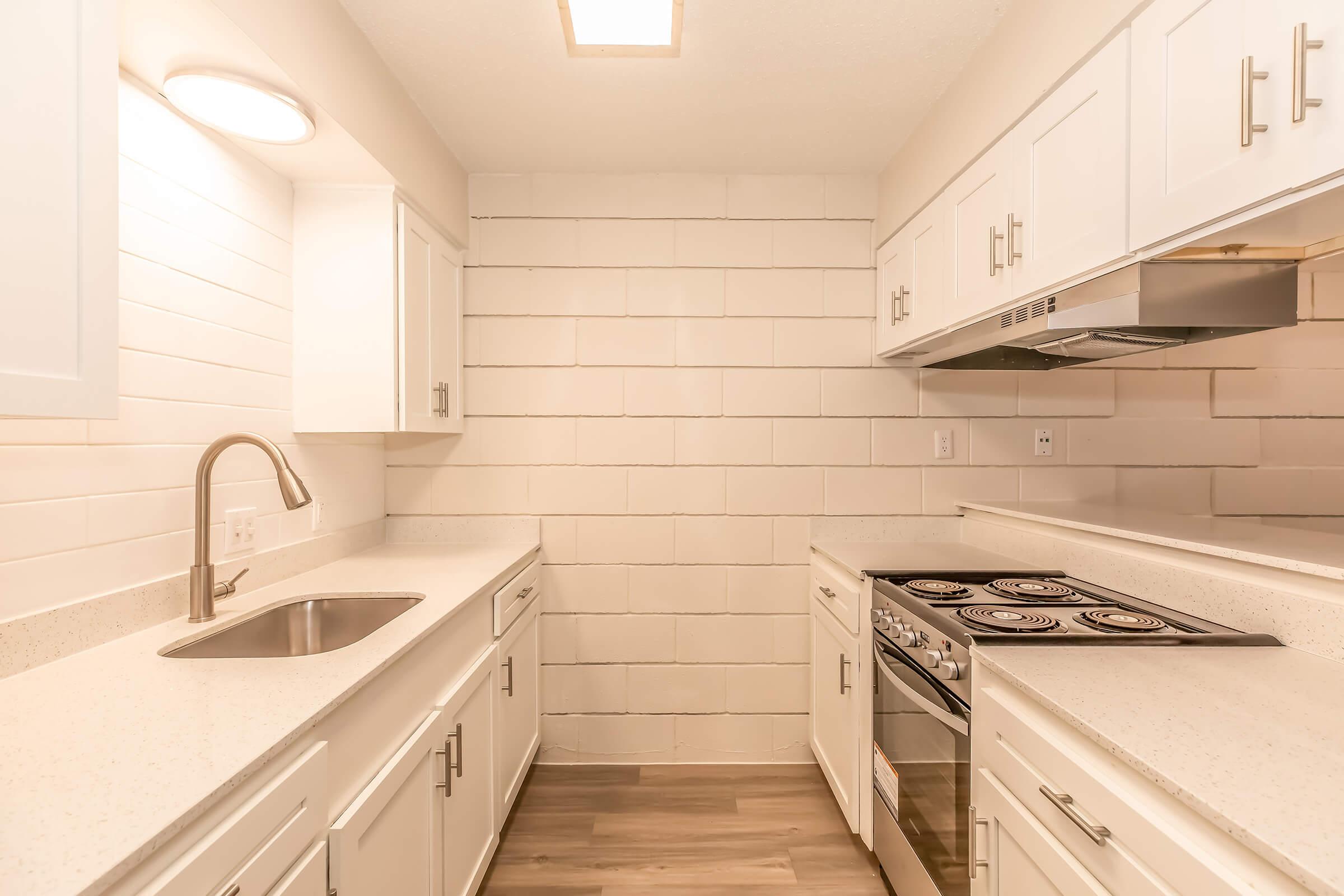 Modern kitchen with white cabinetry and light-colored countertops. Features a stainless steel sink, stove with four burners, and a vent hood. The walls are covered in white tiles, and there is ambient lighting from a ceiling fixture, creating a bright and clean appearance.
