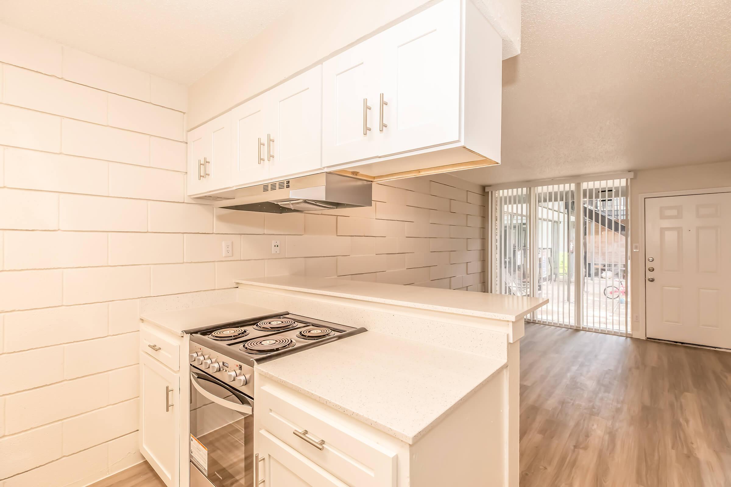 A modern kitchen featuring white cabinetry, a countertop with a built-in stove, and an exhaust hood. The walls are white, and there is natural light from a large sliding door leading to an outdoor space. The floor is light-colored wood laminate.