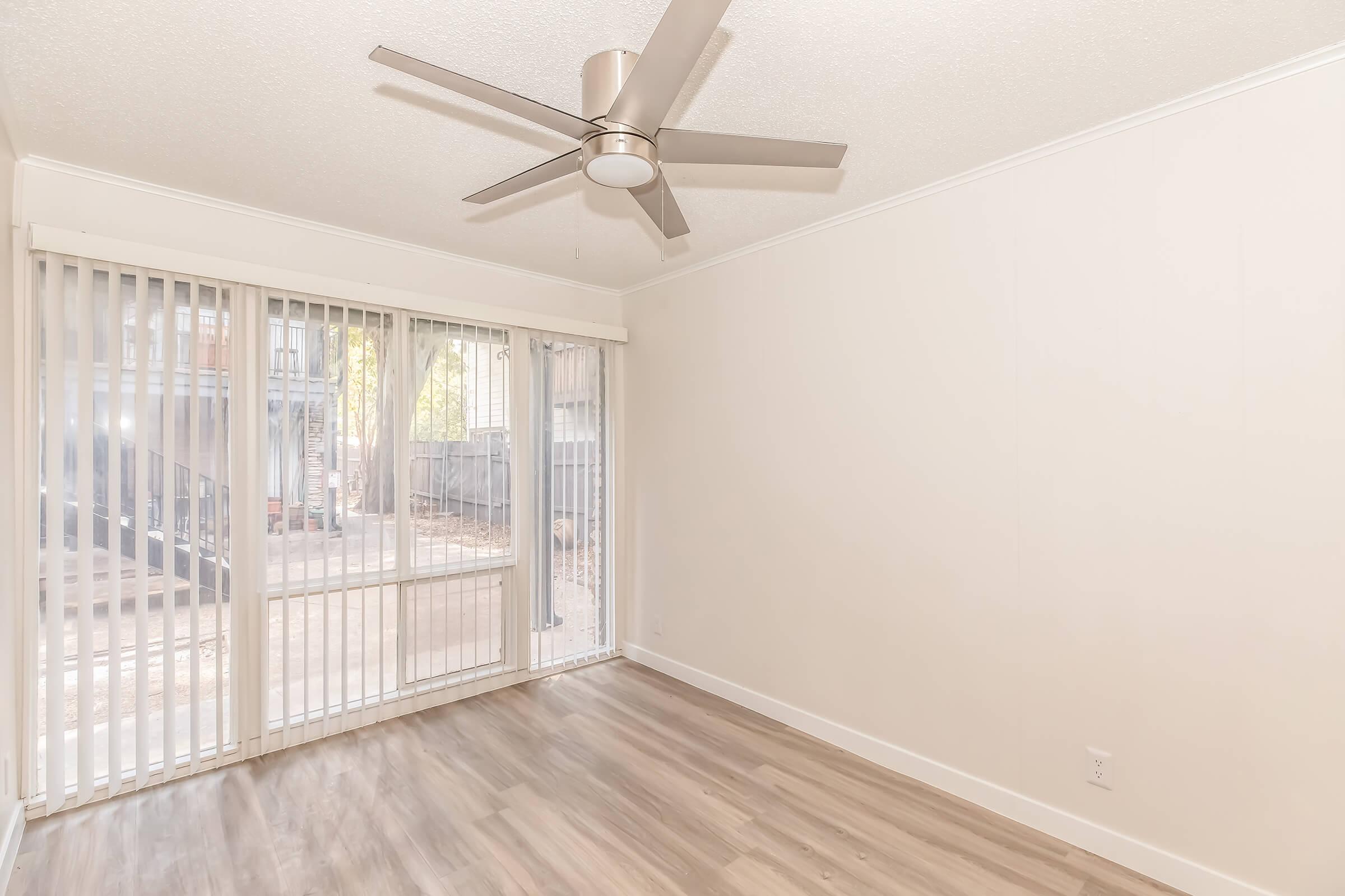 A bright, empty room featuring light wood flooring and large windows with vertical blinds. A modern ceiling fan is centered in the room, which has neutral-colored walls. Natural light streams in, enhancing the spacious feel. The view outside shows trees, adding to the calm atmosphere.