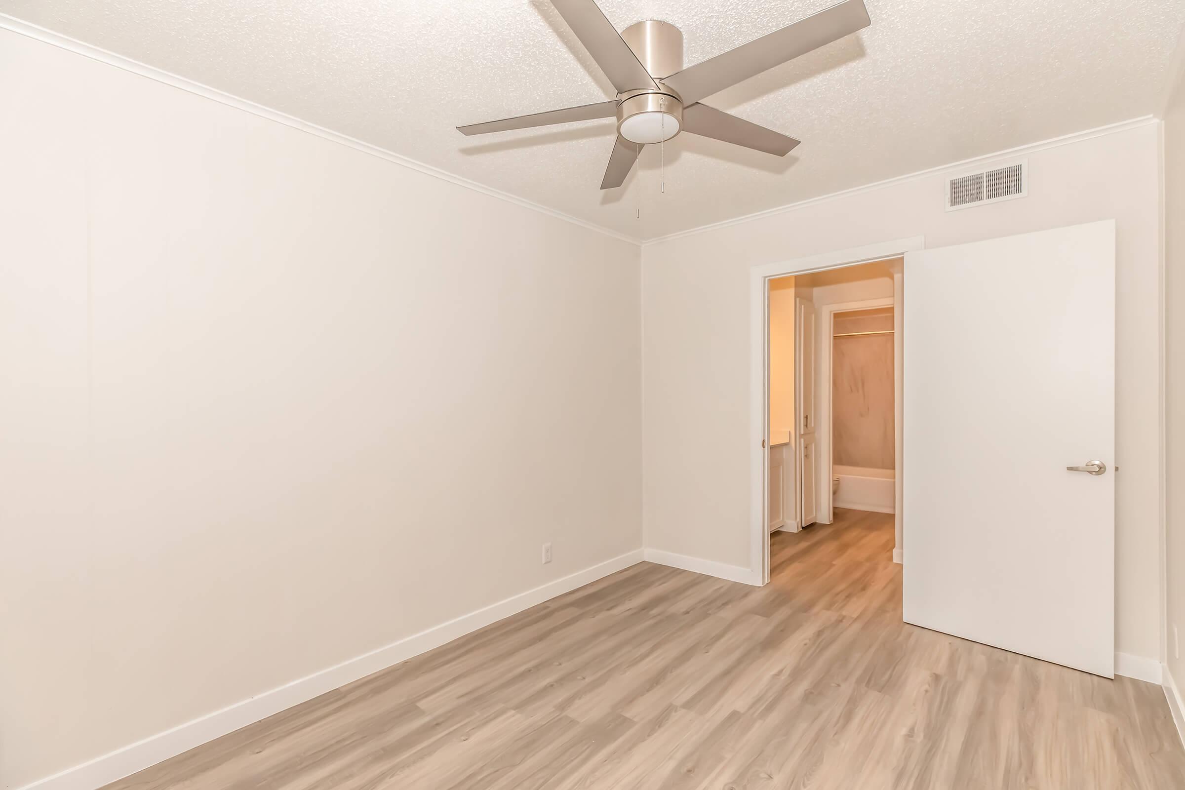 A spacious, empty room with light-colored walls and wooden flooring. A modern ceiling fan is mounted above. A doorway leads to a bathroom area, partially visible in the background. The overall atmosphere is bright and minimalistic.
