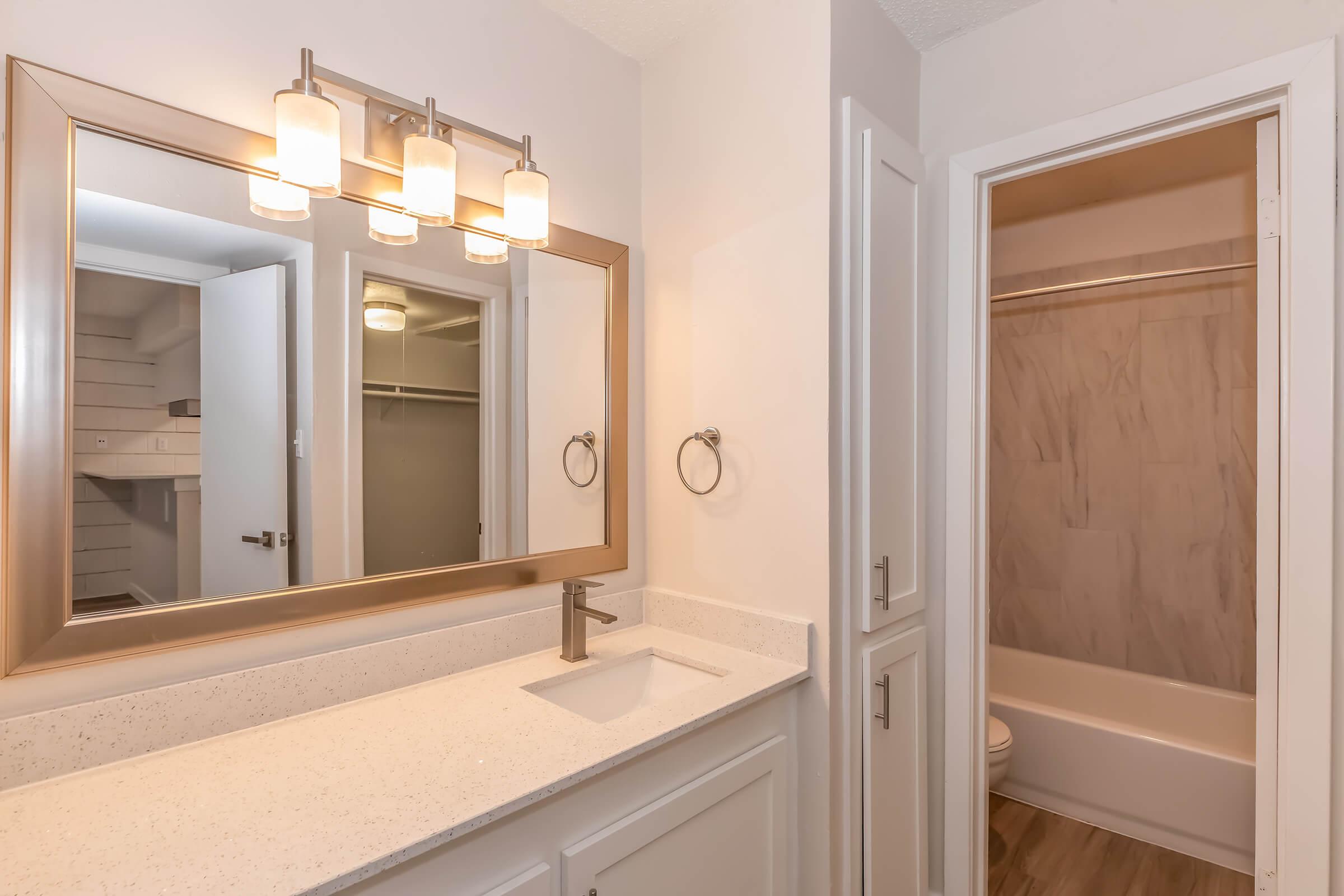 A modern bathroom featuring a white countertop with a sink and a large mirror framed in gold. The lighting consists of three pendant lights above the mirror. To the right, a door leads to a small bathroom area with a bathtub and a toilet, while an open closet with shelves is visible in the background.